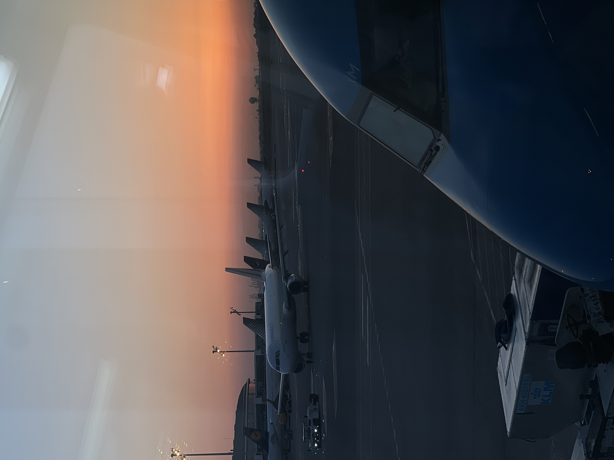 The image depicts an airport tarmac at dusk, viewed through a window. In the foreground, the nose of a blue airplane is visible, with the partial word "XM" on its fuselage. To the left, a cargo container with "KLM" branding is loaded onto a vehicle. In the midground, several airplanes are parked on the tarmac. One prominent white airplane in the center has "AIRFRANCE" written on its side. Other aircraft with visible tail fins are also present. Airport lighting poles with multiple lights are visible in the distance. The background features a building with a curved roof and a city skyline against a sky transitioning from light blue to a vibrant orange sunset. The overall scene suggests operations at an airport, possibly Barcelona-El Prat Airport, located in El Prat de Llobregat, Spain, during the early evening hours.