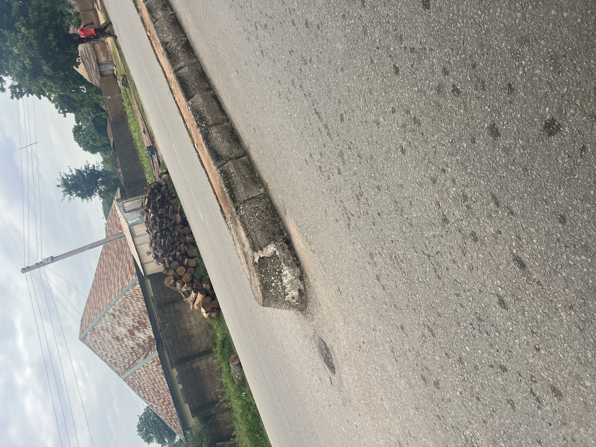 This media file depicts a street scene in Kona Garu, Nigeria. The image is oriented vertically, with the road and its curb taking up a significant portion of the frame. The road surface is asphalt, with visible aggregate and some darker stains. The curb is made of concrete blocks and appears weathered.
To the left of the road, there is a grassy area and a cluster of cut logs, suggesting a recent logging activity or firewood preparation. Further to the left, a building with a corrugated metal roof, showing signs of rust, is visible. Adjacent to this building, there's a smaller structure with a thatched roof. A person wearing a red shirt is standing near the thatched-roof building. Power lines and a wooden utility pole are also present, indicating the presence of electricity. The sky is overcast, suggesting cloudy weather, and the lighting is diffused, implying it might be daytime but not necessarily bright sunlight. The overall scene conveys a rural or semi-rural atmosphere typical of some areas in Nigeria. No visible text is present in the image.