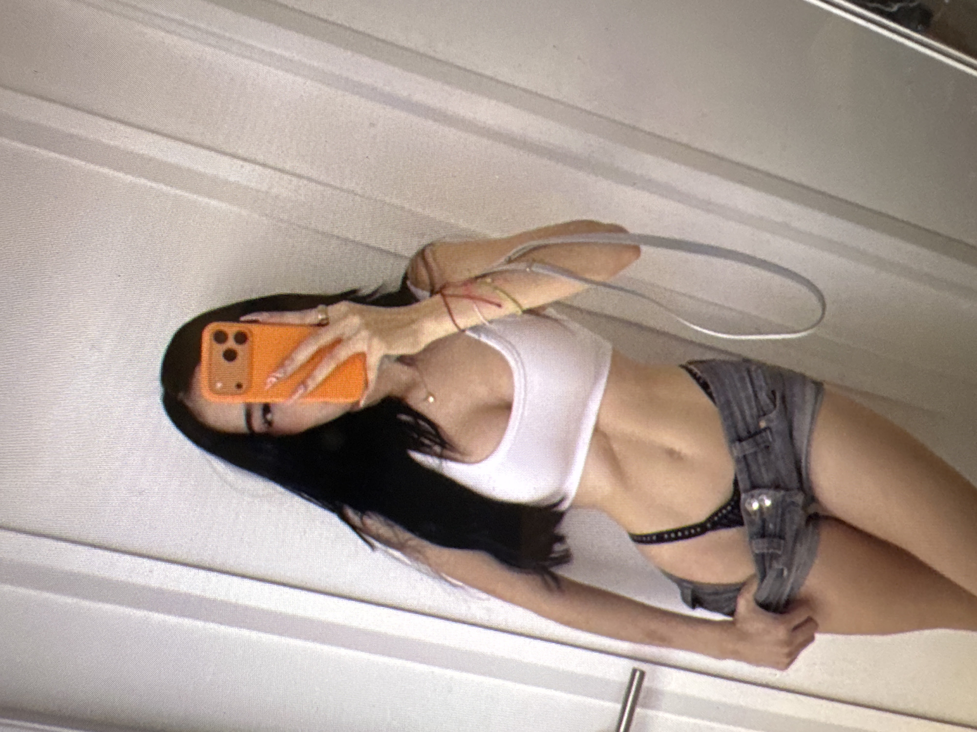 A person with long dark hair is taking a selfie in a room. They are wearing a white crop top, denim shorts, and black underwear. The person is holding an orange phone in their right hand, covering their face. They are also wearing a ring on their left hand and several bracelets on their right wrist. The background appears to be a wall with vertical paneling. There is a silver door handle on the left side of the image. The image is a close-up shot, focusing on the person's torso and upper body.