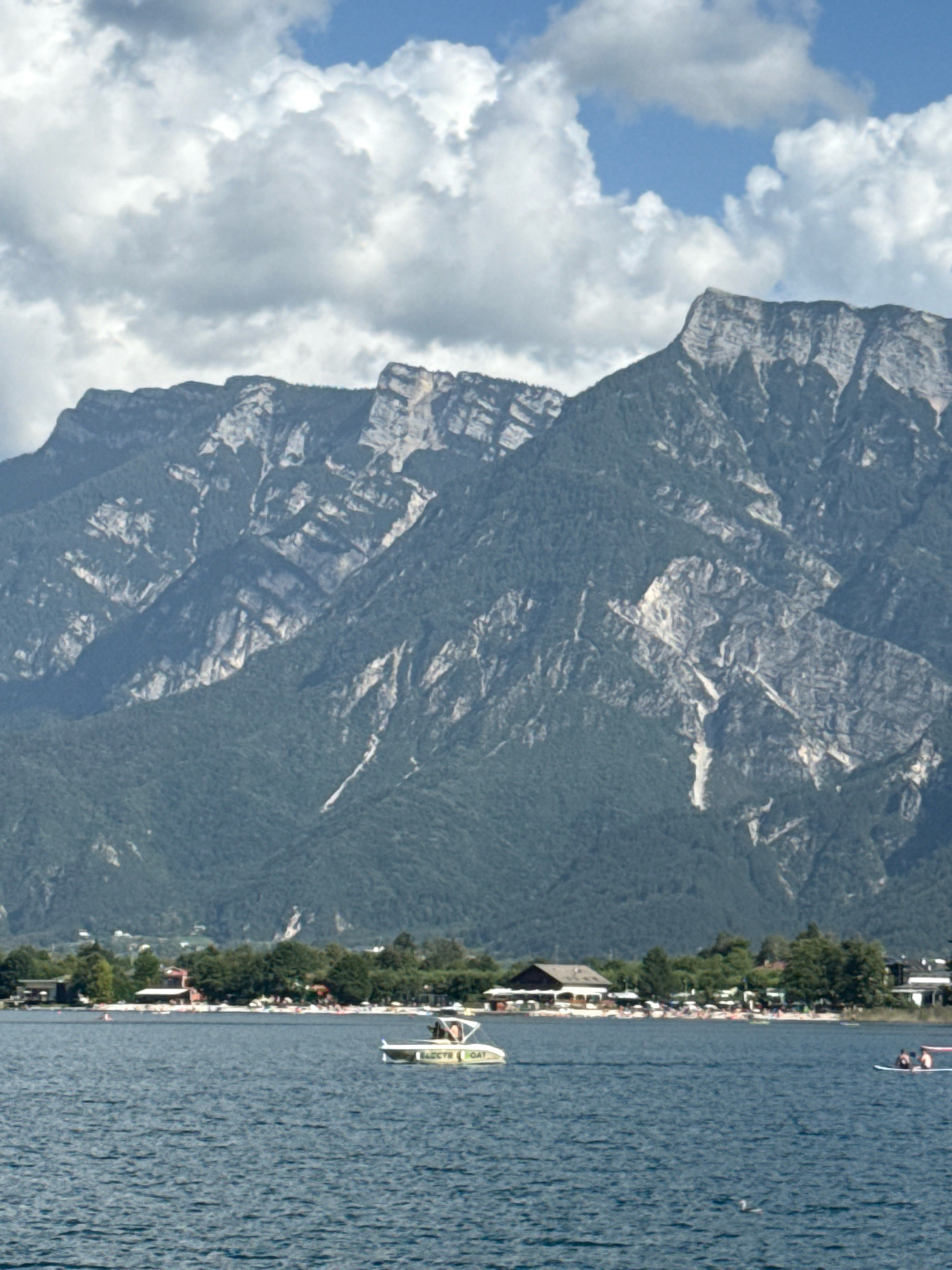 The image depicts a wide shot of the Calceranica al Lago in Italy, featuring a serene lake with a mountainous backdrop. In the foreground, the deep blue water is calm, with gentle ripples distorting the reflections of the sky. A white electric boat with yellow lettering, labeled "ELECTR BOAT," is visible in the middle distance, carrying at least two occupants. Further to the right, two individuals are seated on a paddleboard, enjoying the water.
Along the shoreline, a vibrant scene unfolds. A line of trees separates the water from a beach area, where numerous umbrellas and lounge chairs are set up, suggesting a recreational day. Buildings, possibly restaurants or other lakeside establishments, are interspersed with the trees. The background is dominated by a majestic mountain range, its slopes covered in dark green vegetation, with stark, rocky outcrops and lighter colored cliffs visible at higher elevations. The sky is a mix of blue patches and fluffy white clouds, indicating fair weather. The overall impression is one of a picturesque and tranquil lakeside setting.