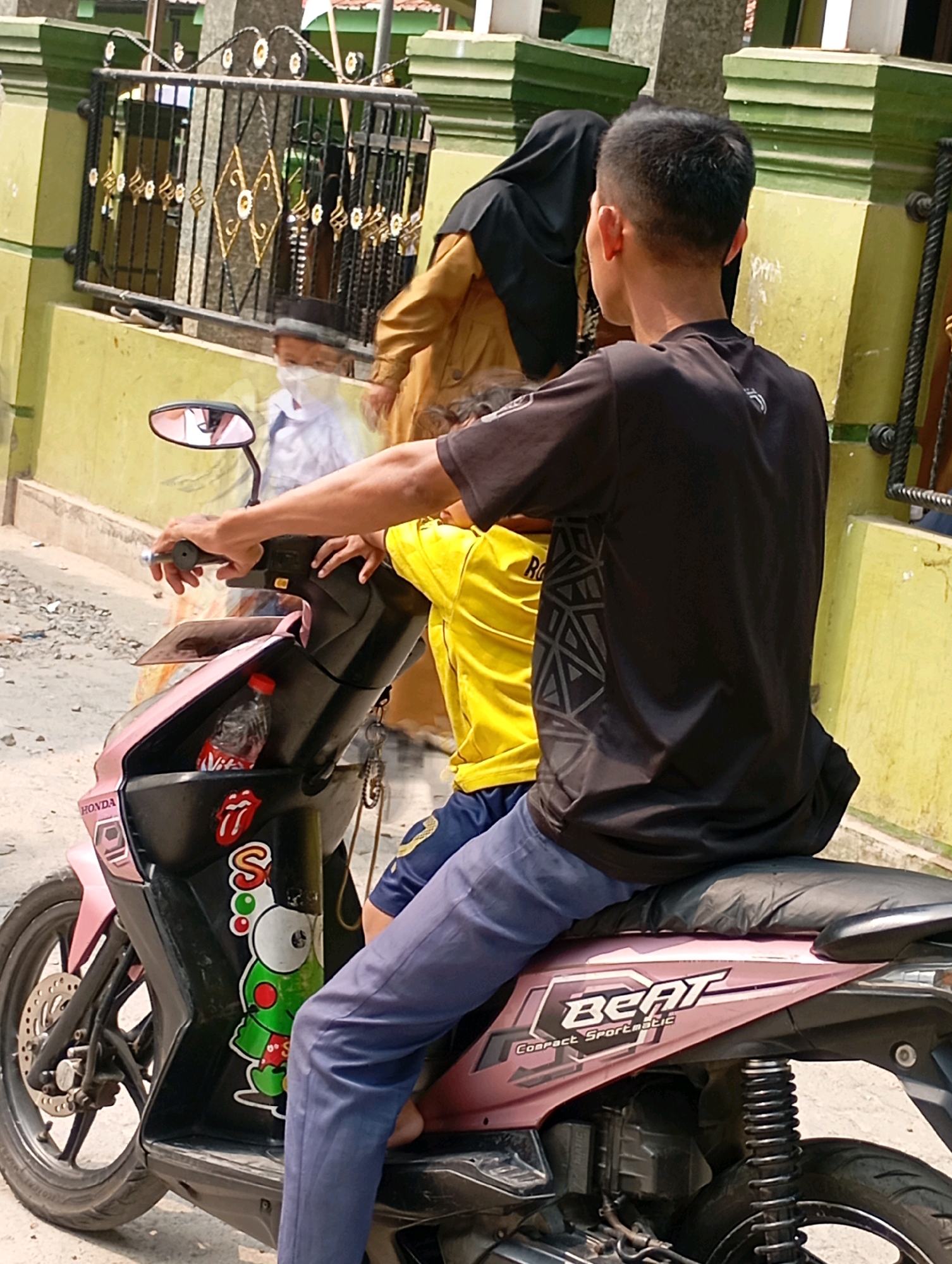 In Babelan, Indonesia, a young male wearing a black t-shirt and blue pants is riding a pink and black scooter. Another individual, partially visible, wearing a yellow t-shirt and blue shorts, is seated behind him. In front of the rider, a young boy in a white shirt, black peci, and face mask is seated on the scooter, partially obscured by the rider's arm. The scooter is parked on a dirt and gravel road, with a yellow building and a black metal fence visible in the background. The scooter has stickers on its front panel, including a Rolling Stones logo and a cartoon frog. A bottle of water is also stored in the scooter's compartment.
