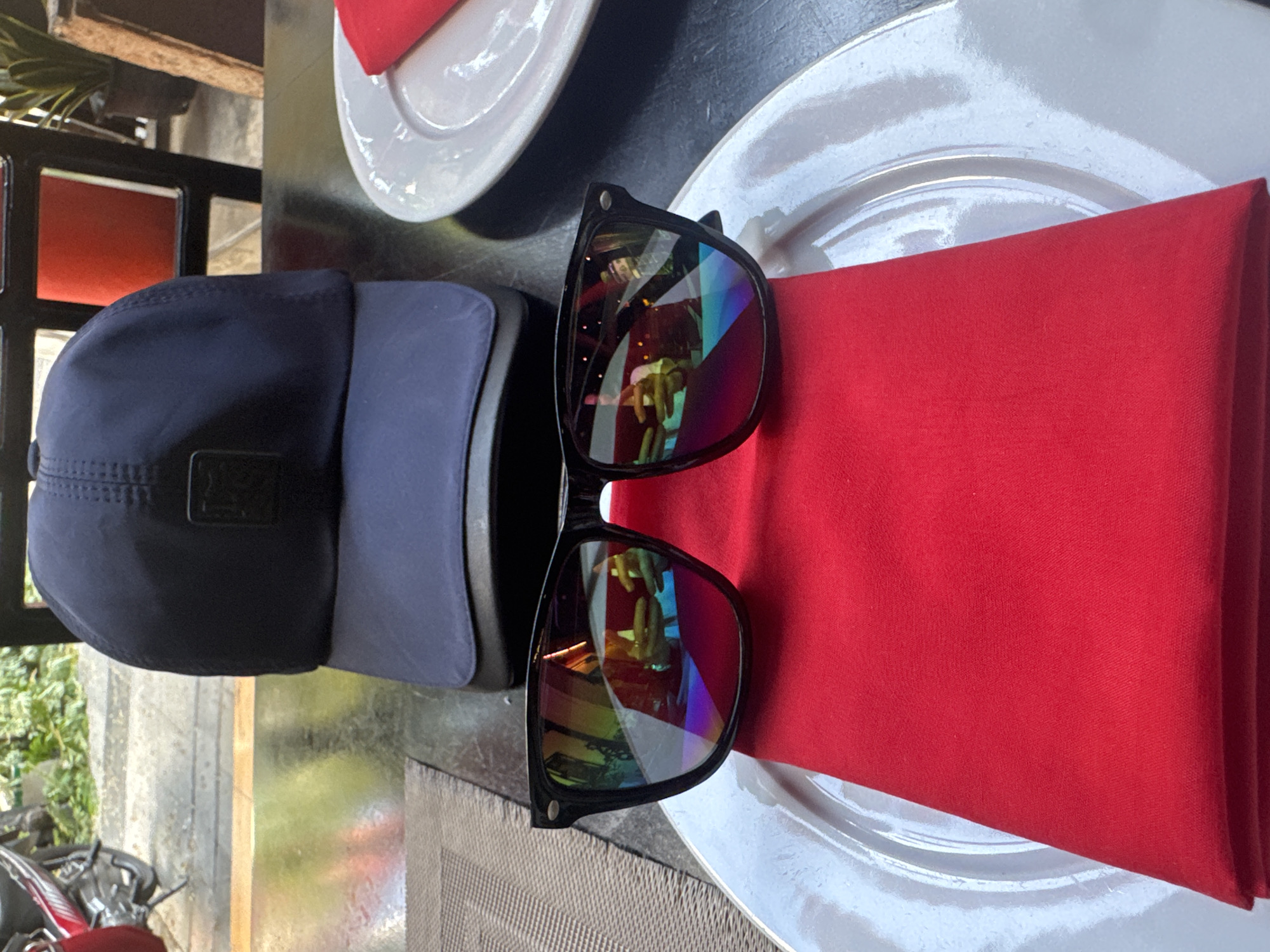 The image captures an overhead view of a table setup, likely outdoors, featuring personal accessories alongside dining items. In the foreground, two baseball caps are stacked, one navy blue on top of a slightly lighter blue or gray one. Next to them, a pair of black-framed sunglasses with highly reflective, iridescent lenses rests on a folded red napkin. The reflection in the sunglasses shows a distorted image of a person's torso and hands, possibly wearing a dark jacket with gold buttons, suggesting a human presence nearby.
The sunglasses and napkin are placed on a white ceramic plate, which is part of a place setting. Another white plate with a red napkin is visible behind the first, further to the top of the frame. The table surface is dark and appears smooth.
The background shows elements suggesting an outdoor setting. To the left, there's green foliage, and parts of a red and black motorbike or scooter are visible. A metal railing or grate structure is also seen in the mid-ground. The lighting suggests it is daytime, with bright reflections on the sunglasses and plates. There is no visible text in the image.