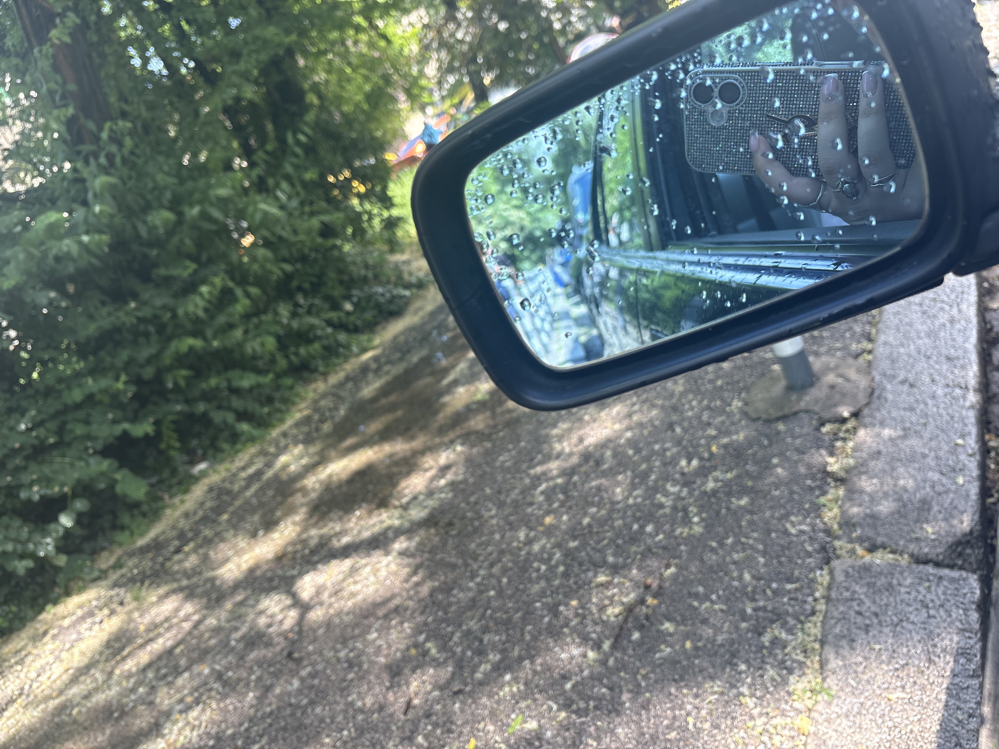 The image captures a close-up view of a car's side mirror, heavily covered in numerous water droplets. The mirror reflects a person's hand holding a smartphone in a distinctive, sparkly, rhinestone-studded case. The hand, adorned with several rings on multiple fingers, features light pink painted fingernails. One ring, on the index finger, appears to have a recognizable emblem or symbol, though it's not clearly discernible.
Outside the mirror, dominating the left and upper portions of the frame, is a dense expanse of lush green foliage, likely trees or bushes, dappled with sunlight. Below this foliage, a textured, light-colored paved or asphalt surface is visible, covered with scattered small, pale-yellow debris, possibly fallen leaves or flower petals. Streaks of sunlight and shadows pattern this ground. On the far right, a gray concrete curb borders the paved area. The bright lighting and shadows indicate it is daytime. The water droplets on the mirror suggest recent rain or a wet environment. No legible text is visible in the image.