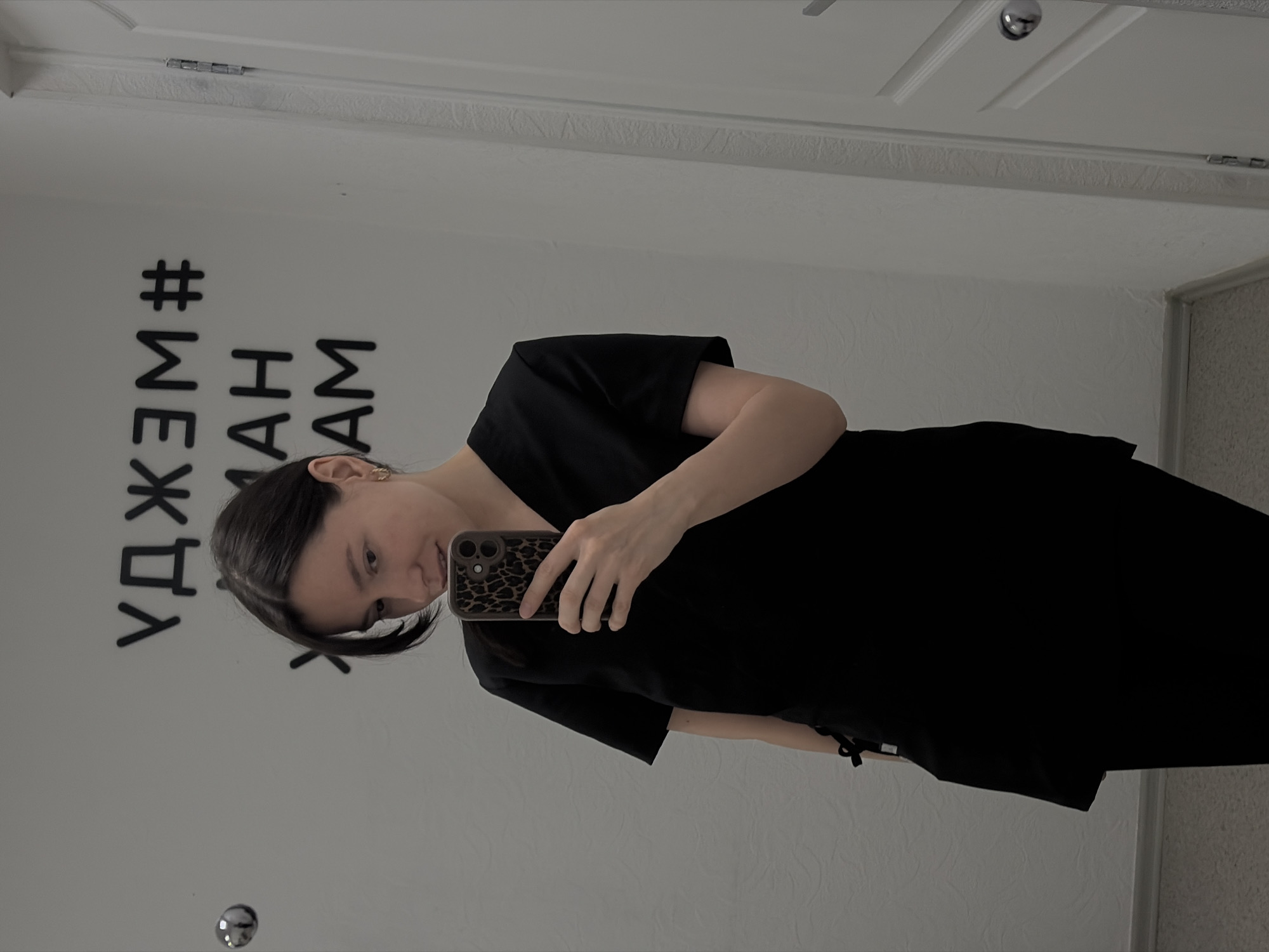A woman is taking a selfie in a mirror. The text "#МЫЖДЕМАМ" is seen on the wall. The image appears to be taken in a room, with a door visible in the background. Based on the text, it is possible that the location is in the city of Chaikovsky, Russia.
