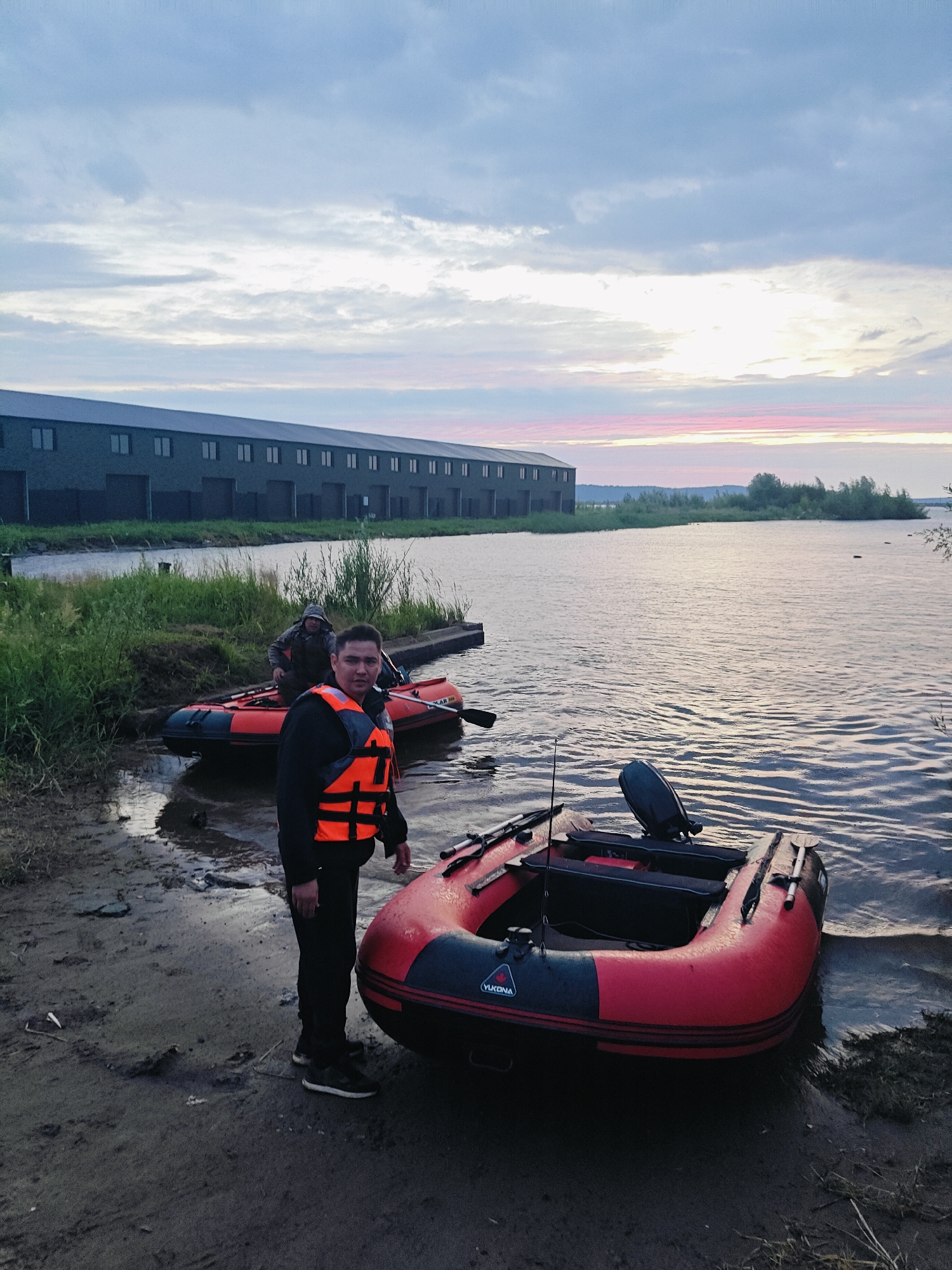 An adult male in an orange life vest and dark clothing stands on a muddy shore next to a red and black inflatable boat, looking towards the camera. In the background, another person is seated in a similar inflatable boat near a wooden dock. The boats are on a body of water with a shoreline of tall grass and a large, multi-windowed building in the distance. The sky is overcast with hints of pink and orange from a sunset or sunrise. The location appears to be a waterfront area in the city of Chaikovsky, Russia.