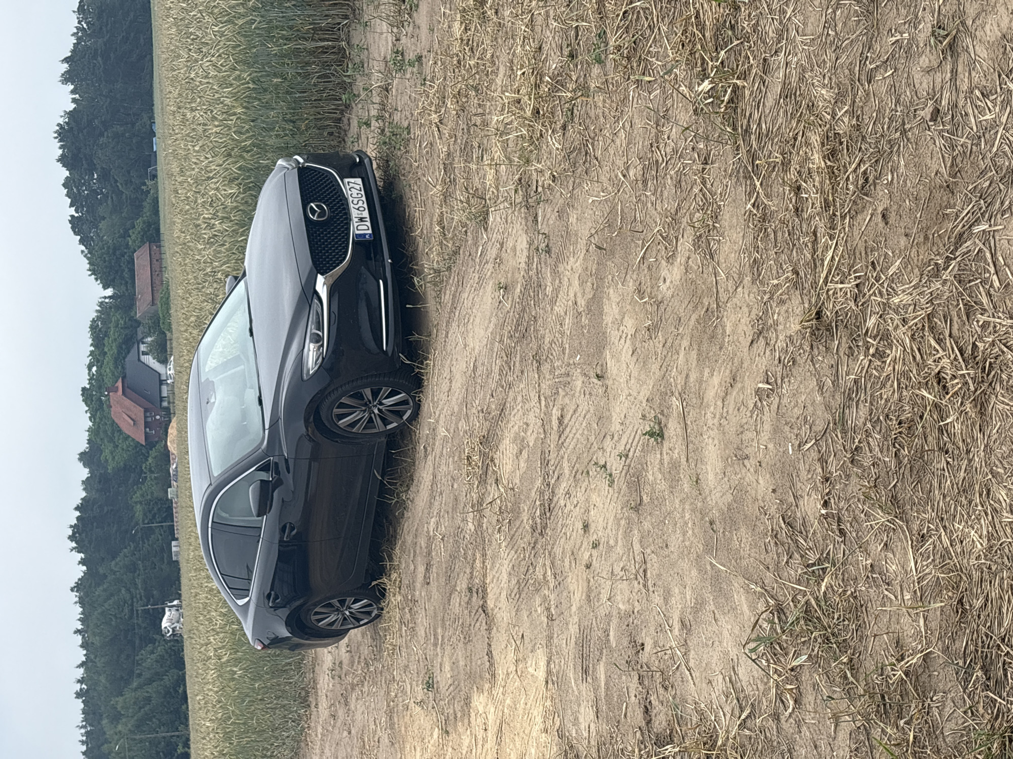 A black Mazda 6 sedan is parked on a dirt road, angled towards the viewer. The car is positioned in the foreground, with its front end slightly to the right. Behind the car, a field of dry, yellowing crops, likely wheat or barley, stretches into the background. Beyond the field, a line of dark green trees forms a backdrop. In the distance, several buildings are visible, including houses with red roofs. One building, possibly a cement mixer truck, is partially obscured by trees. The sky is overcast and appears to be a dull gray. The license plate of the car reads "DW 6SG27". The road surface shows tire tracks, indicating recent vehicle movement. The overall scene is one of a rural or semi-rural environment. The location is identified as being in the vicinity of Opatów, Poland.