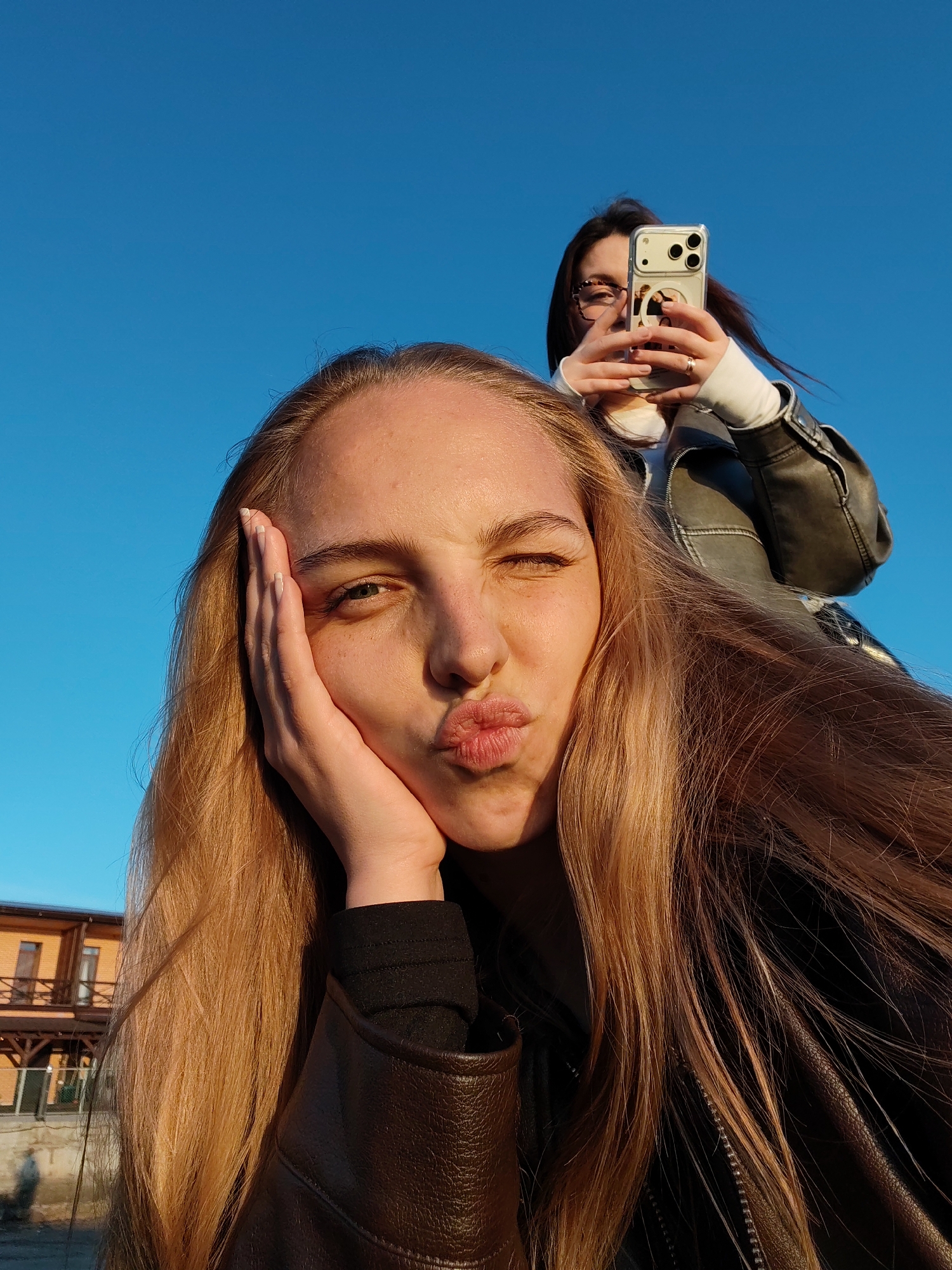 A selfie of a young woman with long blonde hair is being taken outdoors in Chaikovsky, Russia, with another young woman holding a smartphone in the background. The foreground woman is winking and puckering her lips in a kiss, resting her face on her hand. She is wearing a black top and a brown leather jacket. Her hair is golden blonde and styled to fall over her left shoulder. Her skin is fair and lightly freckled.
The background woman is holding a light-colored smartphone with multiple camera lenses, presumably taking the photo. She is wearing glasses, a white sweatshirt, and a leather jacket.
The background features a clear blue sky. To the left, part of a brown brick building is visible. The selfie appears to be taken at ground level, with glimpses of a building and street-level details. The environment suggests a paved or concrete area. The lighting is strong and direct, indicating a sunny day.
