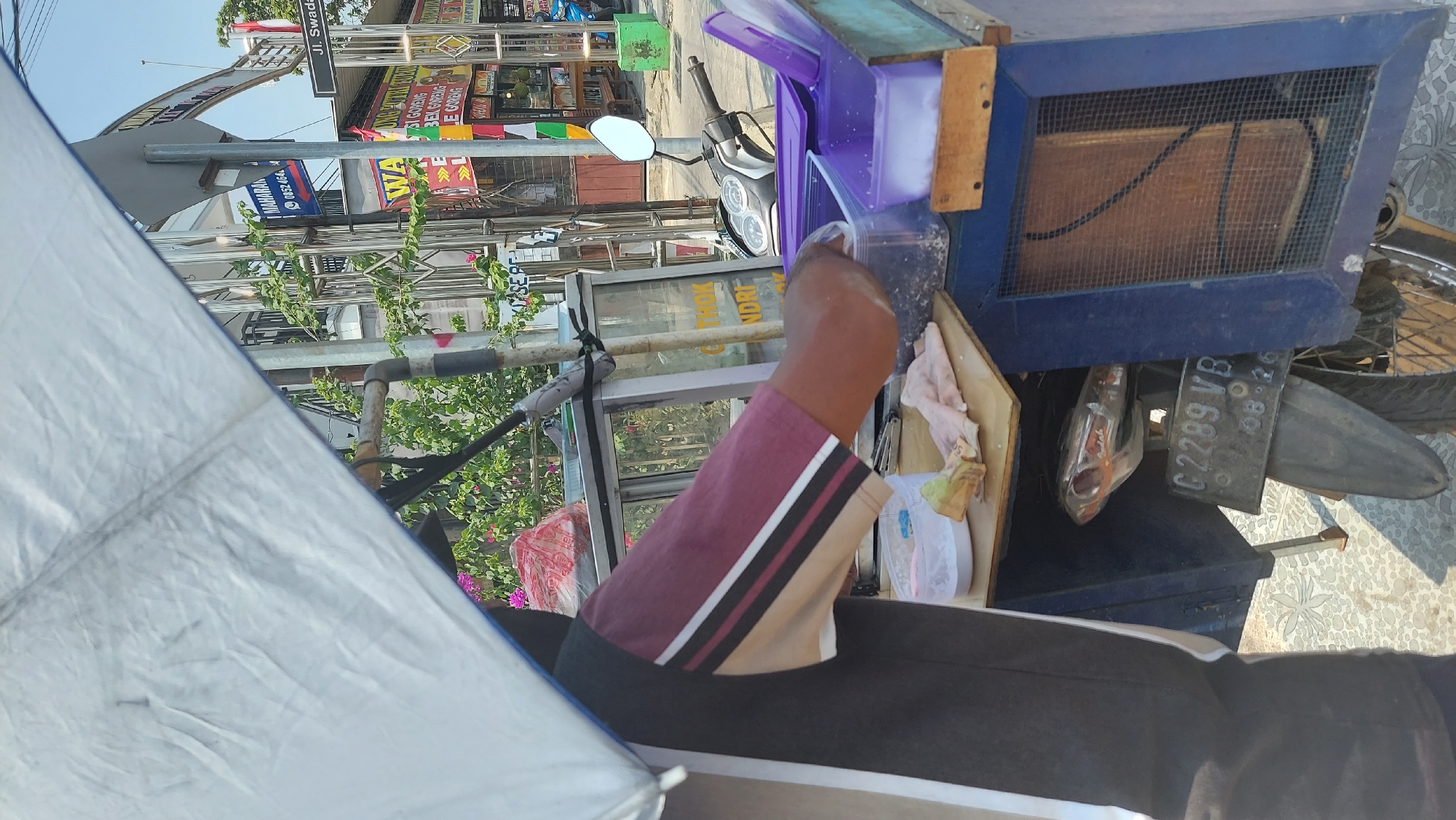 A street vendor prepares food from a cart attached to a motorcycle in Gayam, Indonesia. The cart is covered by a large, white umbrella. A person, wearing a maroon and black shirt, is seen from the waist down, reaching into a plastic container on the cart. The cart holds various containers, some with food items, and a cloth rag is draped over a wooden surface. A motorcycle is parked behind the cart, its license plate reading "C 2289 VB 08-26". In the background, signs for businesses and a street sign with "Jl. Swadaya" are visible. The environment appears to be a sunny street with some vegetation.