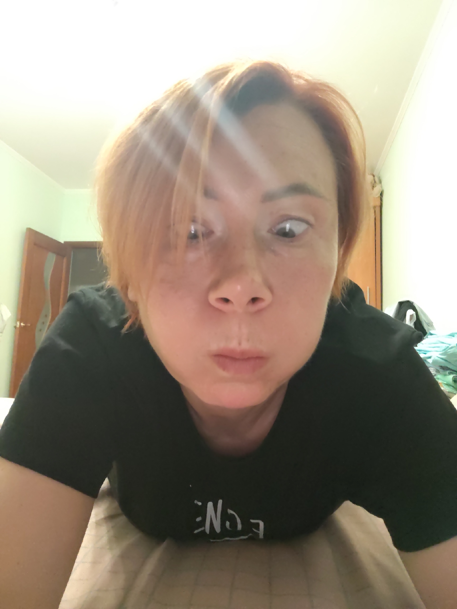 A woman with auburn hair lies on a bed, her face close to the camera. Her eyes are wide and looking in different directions. She is wearing a black t-shirt with white lettering. The room appears to be a bedroom, with a door visible in the background. The walls are painted a light green. The scene is indoors. The woman's location is unknown, but the context suggests it is a residential setting.