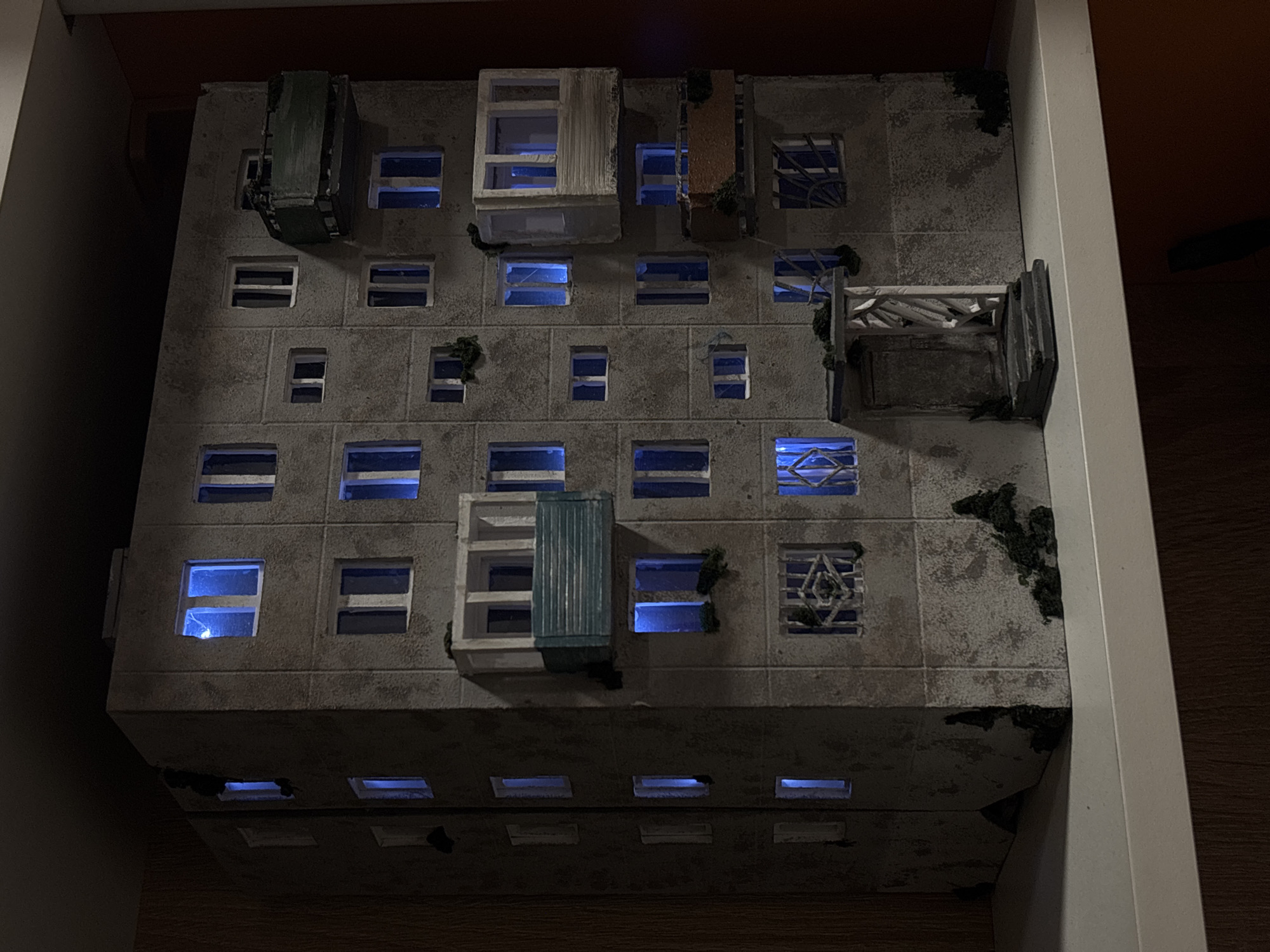 The image displays a miniature model of a dilapidated, brutalist apartment building. The building is gray with dark accents and features multiple rows of windows with a blue light shining from within. Several balconies of varying designs protrude from the facade, some displaying signs of decay with small patches of moss or plant life. The model sits inside a drawer with wooden bottom and white sides. It is difficult to ascertain the specific location within Perm, Russia, based on the model alone. The scene is dimly lit, enhancing the model's somber atmosphere.