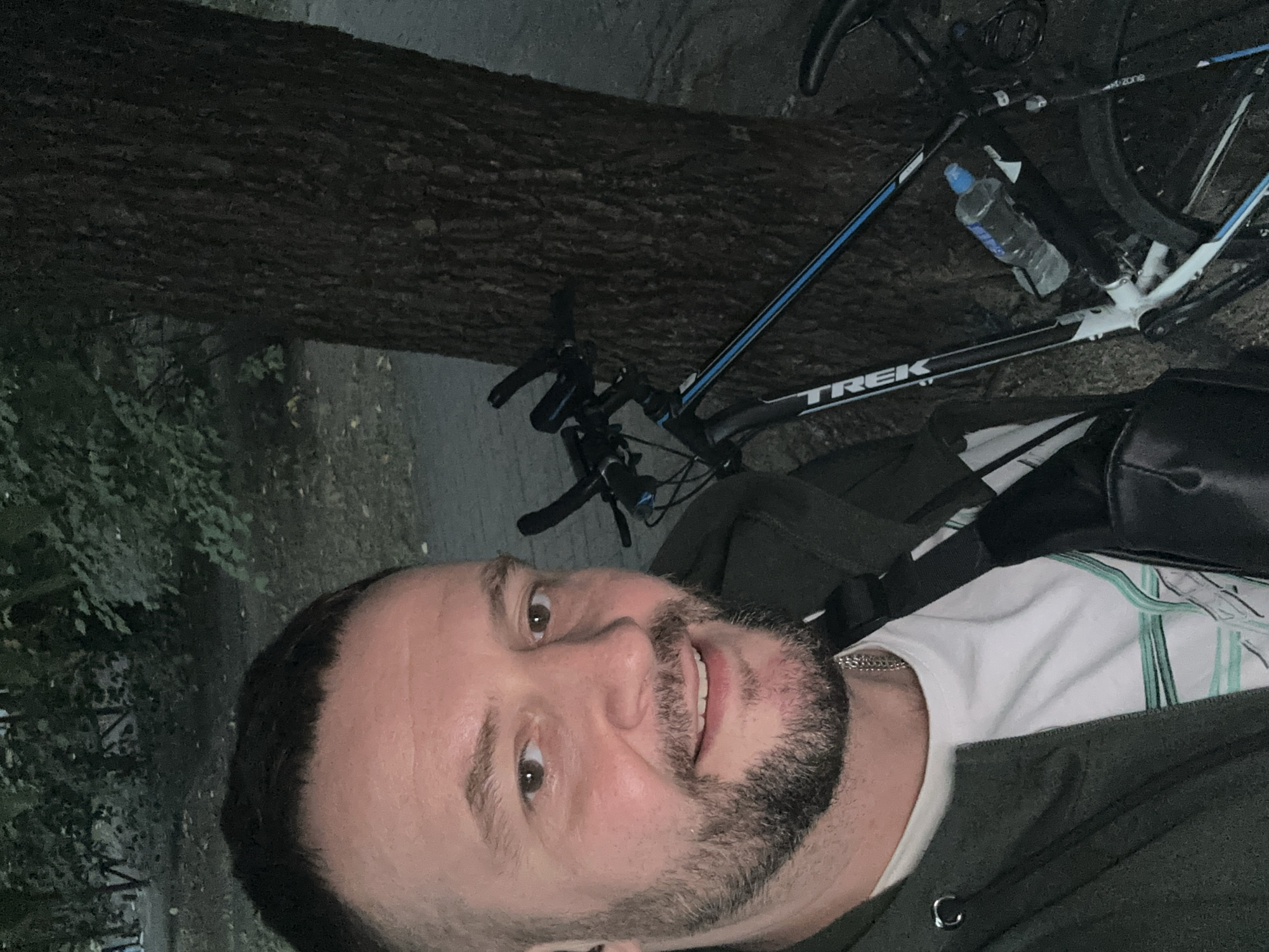 A man with short dark hair and a beard is taking a selfie outdoors, leaning against a tree. He is wearing a dark green hooded jacket over a white t-shirt with a striped pattern. A black crossbody bag is slung across his chest. A Trek bicycle with a water bottle attached to the frame is parked next to him. The background shows trees and a paved path, suggesting a park or outdoor recreational area. The overall lighting indicates it is either early morning or late evening. This image was captured in Minsk, Belarus.