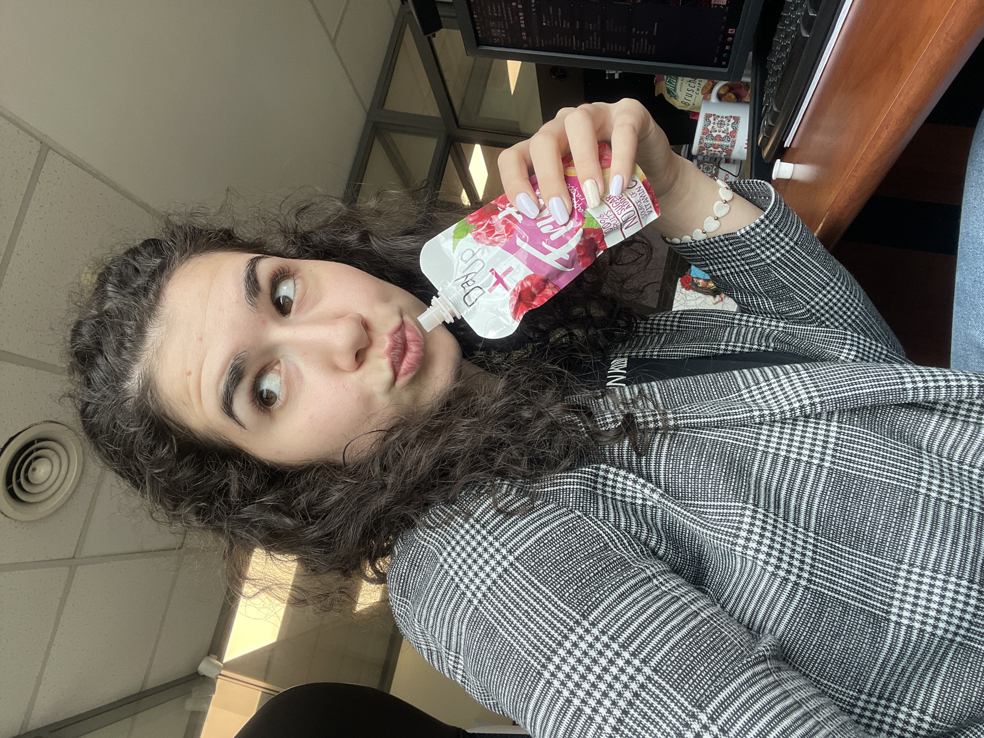 A young woman with curly dark hair is drinking from a raspberry-flavored fruit puree pouch labeled "Day Up." She is wearing a black and white plaid blazer over a black shirt, with a white heart bracelet on her right wrist. Her nails are painted with a light pastel lilac. The background shows an office setting, including a desk, computer monitor, and a mug with traditional floral patterns. The scene is well-lit, with natural light coming in from a window, and appears to be indoors. Based on the cup in the background reading "Ma Brusch," it is likely located in Sofia, Bulgaria.