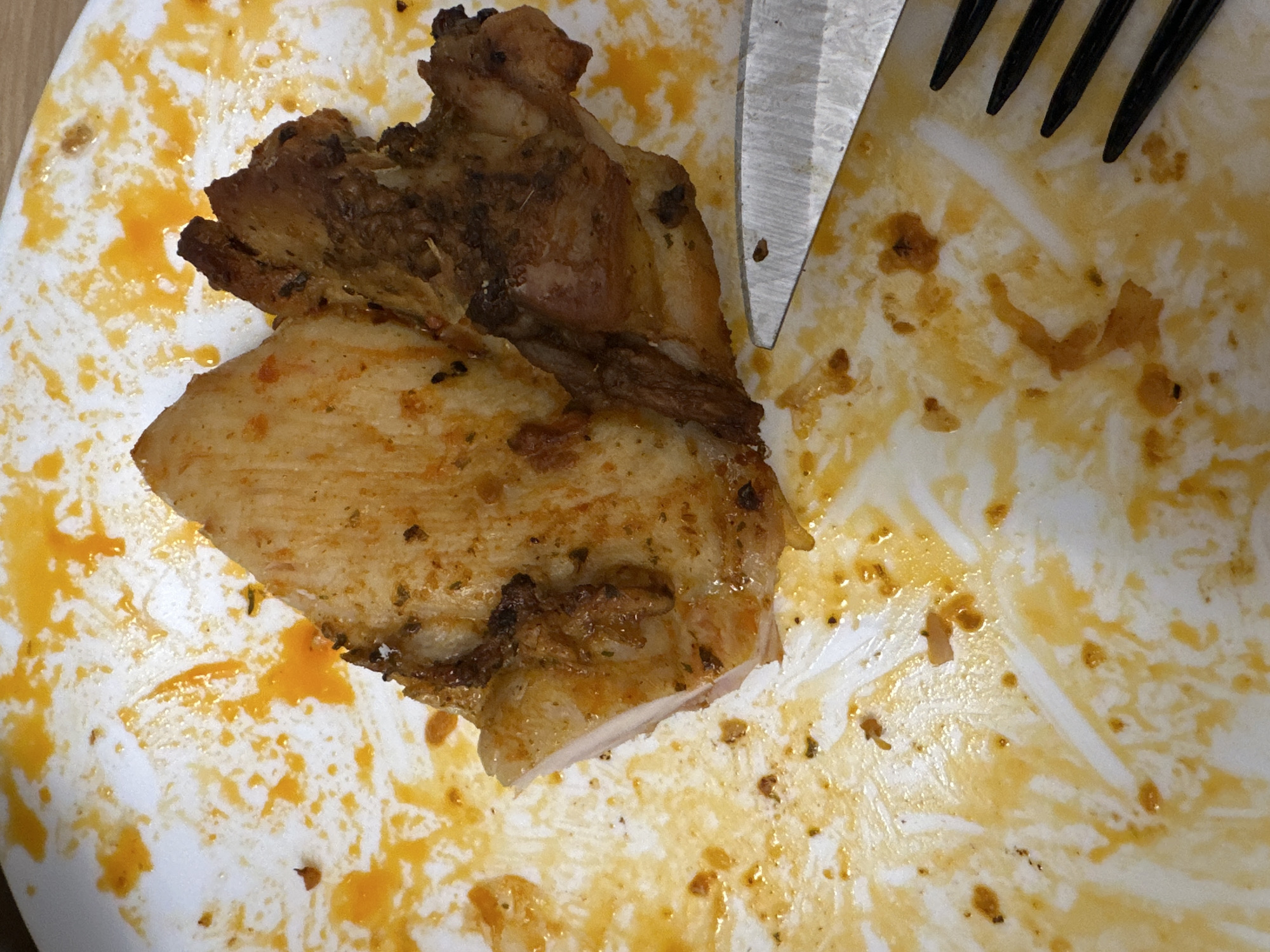 A close-up, high-angle shot captures a partially eaten meal on a white ceramic plate, set in Sofia, Bulgaria. At the center-left, a single piece of cooked chicken, likely a portion of a thigh or similar cut, rests on the plate. The chicken exhibits a reddish-orange seasoning, suggesting spices and herbs, and one edge reveals a clean cut, showing the tender, white cooked meat within. A darker, more textured section, possibly skin or bone, is visible on the upper part of the chicken.
The white plate is heavily stained with glossy, orange-yellow grease and sauce remnants, along with scattered small dark specks, indicating a flavorful and well-seasoned dish. A silver-colored knife is visible on the right side of the frame, its blade pointing towards the chicken, while the black prongs of a fork are partially visible in the upper right corner, suggesting that the meal is actively being consumed or has just concluded. No people are visible in the image, and the specific time of day or external weather conditions cannot be determined from this indoor setting. There is no visible text.