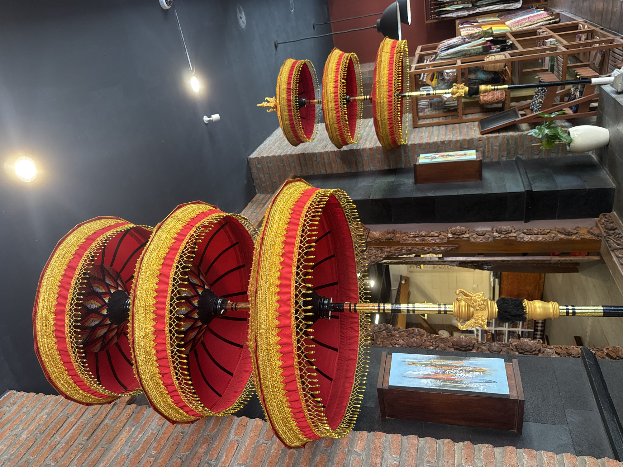 The image shows an indoor display of traditional Balinese umbrellas. Three large, red and gold umbrellas are suspended at the bottom of the frame, while three smaller versions are visible in the background. The umbrellas feature ornate gold trim and are attached to a golden shaft. They are displayed in a building with black walls, brick accents, and black tiled flooring. In the background, there is a view to another level of the building where fabrics are visible on shelves. The location is likely a shop or gallery in Kuta Selatan, Indonesia, based on the presence of Balinese umbrellas, which are cultural symbols of the region. A painting in a wooden frame rests on a surface. The lighting appears to be a combination of artificial lighting and natural light.