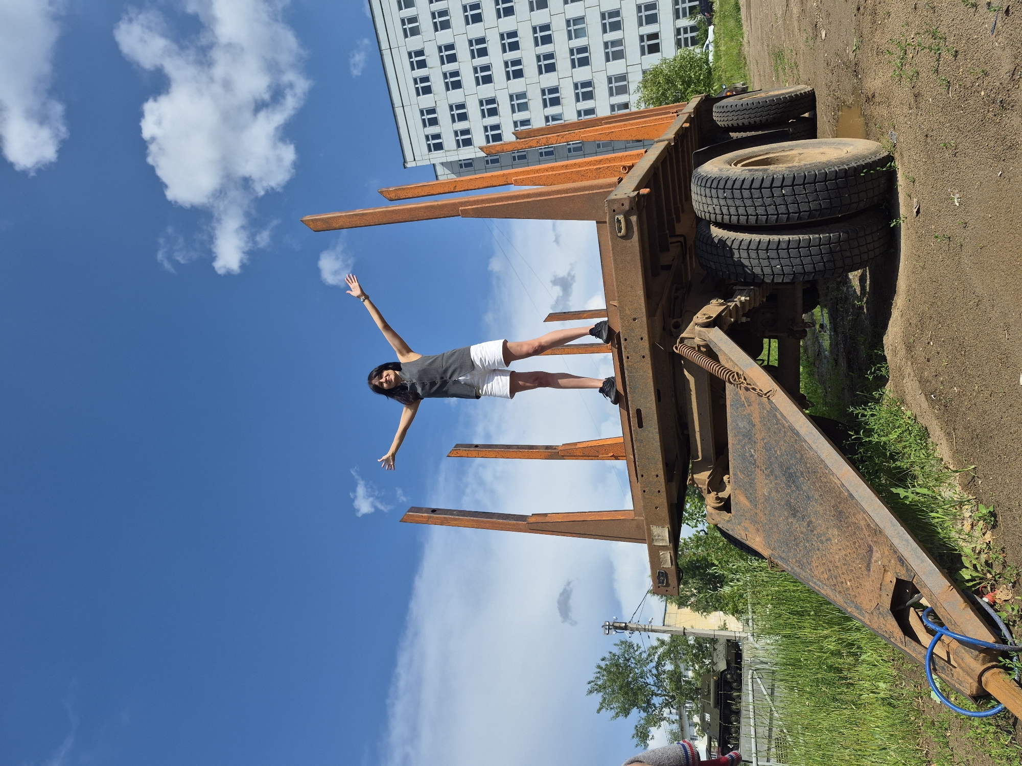 In Chaikovsky, Russia, on a sunny day with a bright blue sky and scattered clouds, a young woman stands on an empty, rusty orange trailer. She wears a dark sleeveless vest over a white t-shirt and white shorts, with black sneakers. Her arms are outstretched to her sides, and her head is tilted back slightly. The trailer has tall, vertical wooden stakes along its sides and a ramp at the front. In the background, a multi-story white building with numerous windows is visible. To the left of the trailer, a military vehicle is partially visible, along with green grass and trees. The ground is dirt.