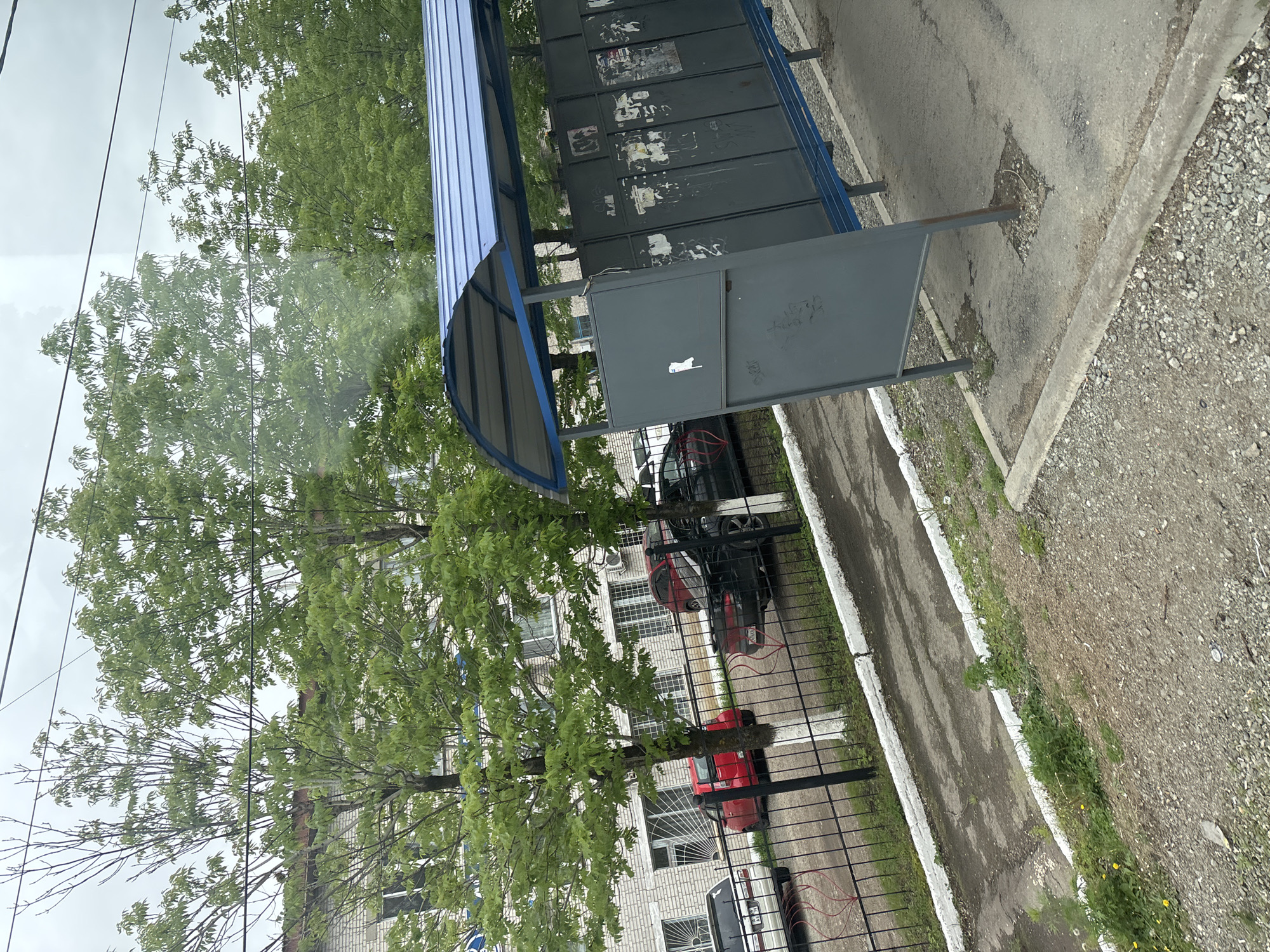 The image displays an outdoor scene, likely an urban or suburban area, viewed from a slightly elevated perspective. In the center, a utilitarian shelter with a curved blue corrugated metal roof and gray metal walls stands on a paved surface. The back panels of the shelter are adorned with patches of torn posters and faded graffiti. A smaller, dark gray metal box or panel is attached to the side of the shelter, exhibiting faint scribbles, including a stylized white outline that resembles a cat.
Behind the shelter and to the left, several trees with abundant green foliage partially obscure a multi-story building made of light-colored brick. The building features numerous windows, some of which have security grilles. A black metal fence runs along the perimeter of the building's property, behind which several cars are parked; a black car, a red car, and part of a light-colored vehicle are visible.
The ground in the foreground and to the right consists of a worn paved area (likely asphalt or concrete), a concrete curb, and a gravel-covered section. Patches of green grass and some small yellow flowers grow along the edges of the paved areas. The sky overhead is overcast and cloudy, with several thin power lines traversing it. The natural light indicates it is daytime, but there are no harsh shadows. No people are present, and the scene depicts a quiet, static moment in an everyday environment.