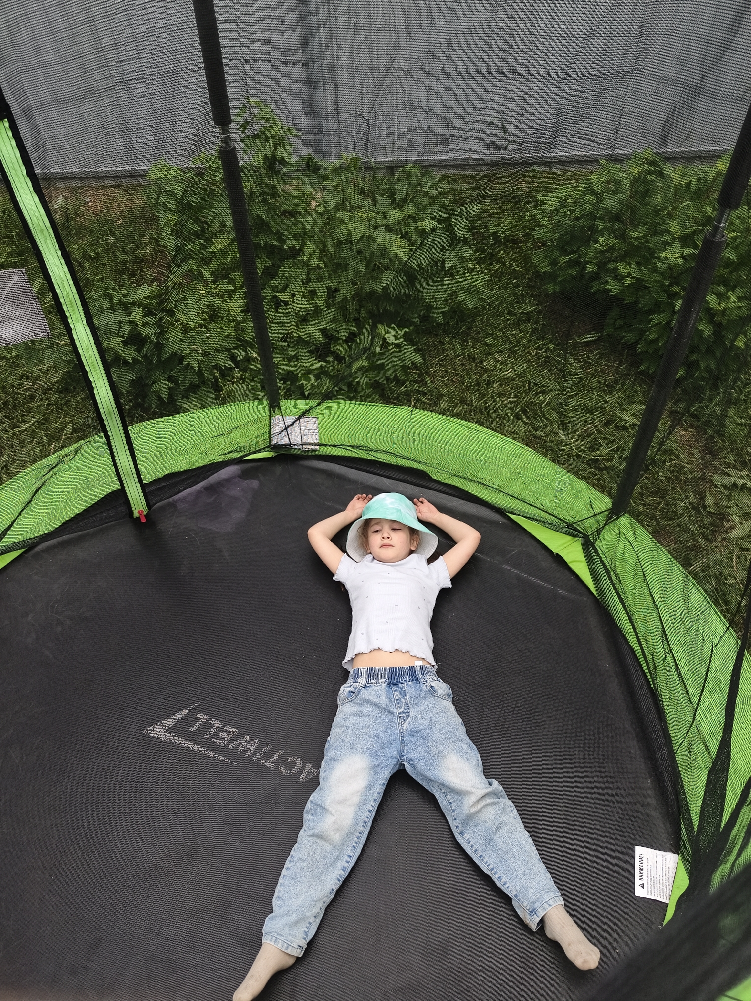 An overhead view depicts a young girl lying supine on a black outdoor trampoline in Prikamskii, Russia. The girl, estimated to be between 4 and 7 years old, wears a light blue bucket hat, a white short-sleeved t-shirt with a slightly ruffled hem, light-wash blue jeans, and light grey socks. Her arms are bent with hands resting above her head, and her legs are slightly splayed. Her eyes are open, gazing upwards with a neutral expression.

The trampoline features a black jumping mat with the reversed white "ACTIVWELL" logo and an arrow pointing towards the top of the frame. A bright lime green safety pad covers the trampoline's springs and frame perimeter. A black mesh safety enclosure surrounds the trampoline, supported by four visible black padded vertical poles. A red-tabbed zipper is visible on the green frame cover on the left side, serving as the entrance. A white warning label with partially visible black text, including "ВНИМАНИЕ!" (Russian for "ATTENTION!"), is attached to the green padding near the girl's right leg.

Beyond the trampoline's mesh enclosure, dense, vibrant green foliage, appearing as wild bushes and weeds, covers the ground. Above the vegetation, a dark grey, finely woven mesh structure extends across the upper background, suggesting either a shade cloth or a larger enclosure. The scene is illuminated by bright, natural daylight, indicating an outdoor setting.
