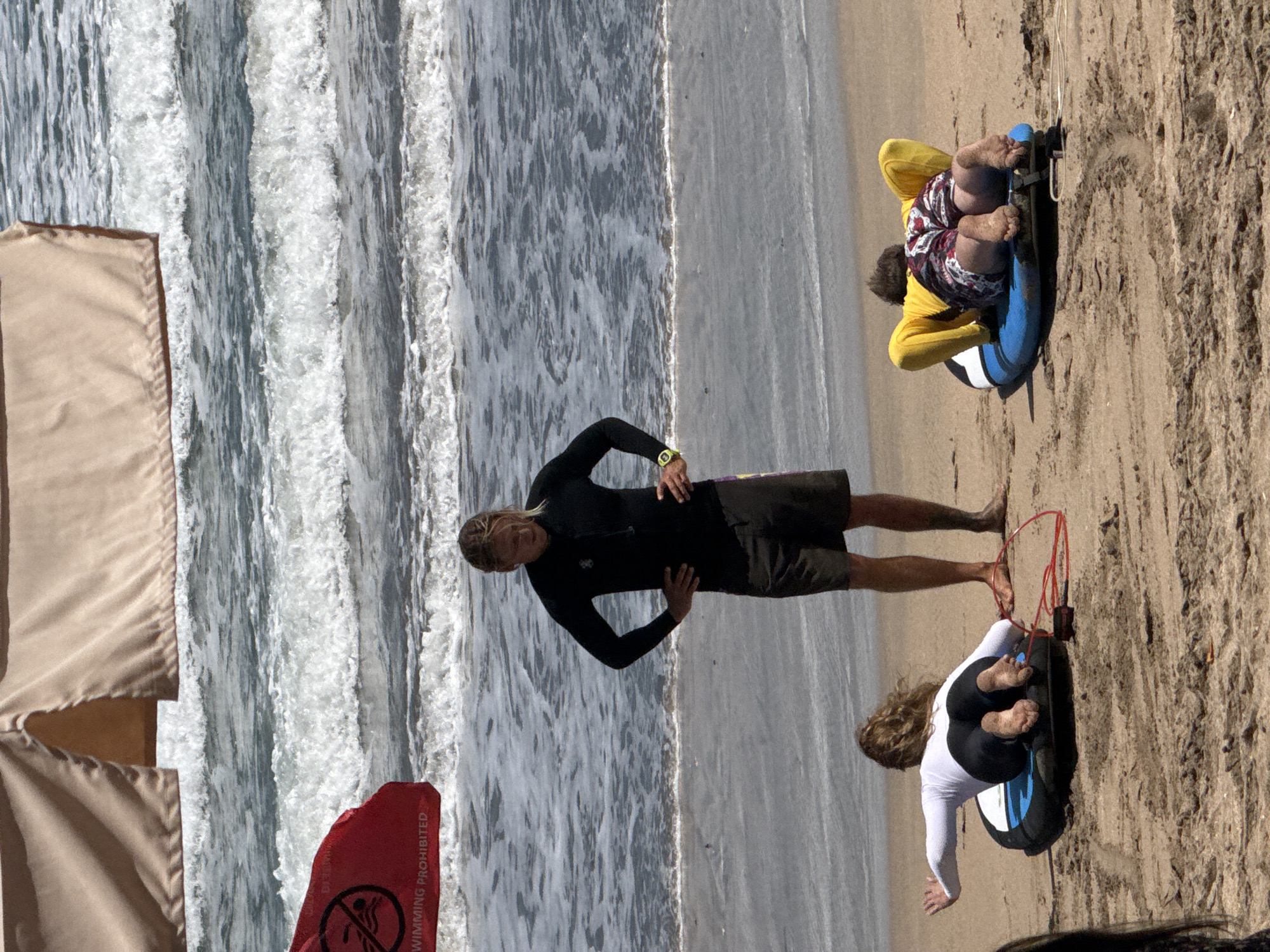 A surfing instructor, a man with blonde hair wearing a black wetsuit and dark shorts, stands on a sandy beach in Kuta, Indonesia, with his hands on his hips, facing the ocean. Two students are lying on their surfboards in front of him. The student on the left is a woman wearing a white long-sleeved shirt and black leggings, her back to the camera. The student on the right is a person wearing a yellow long-sleeved shirt and patterned shorts, also with their back to the camera. The ocean, with white-capped waves, is visible behind them. A red flag with a "no swimming" symbol is planted on the left side of the beach, near the water's edge. A light-colored awning is visible in the upper portion of the image.
