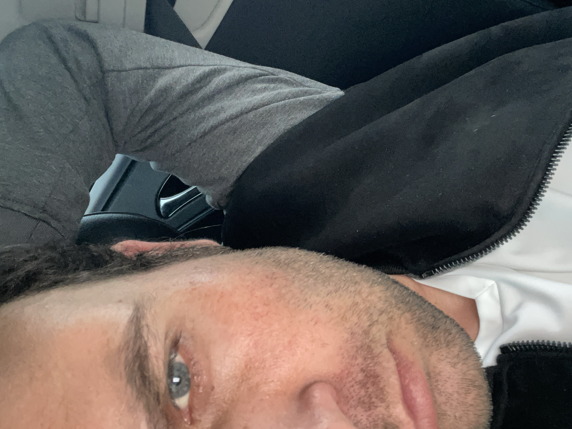 A man is lying down, his head towards the bottom of the frame. He has short dark hair, light skin, and light blue eyes. He is wearing a white shirt with a black jacket over it and grey sweatpants. The background appears to be the interior of a car, with a black seat and part of the steering wheel visible. There is no identifiable location or specific details suggesting this is in the city of Armagh, United Kingdom.