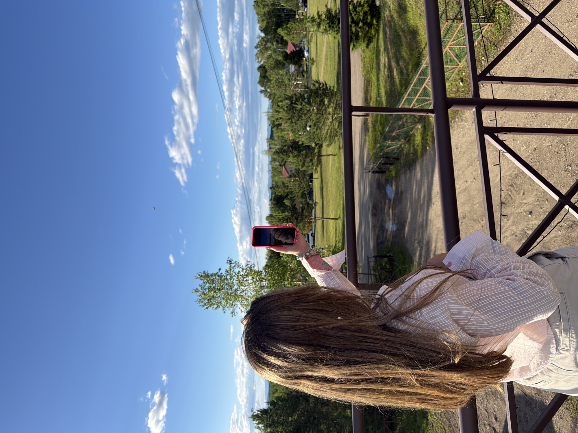 A young woman with long, light brown hair stands on a balcony overlooking a rural landscape. She is holding a pink smartphone in her right hand, seemingly taking a picture or recording video. She is wearing a light pink and white striped shirt and light-colored pants. The background features a grassy field dotted with evergreen trees, houses with red roofs, and a dirt road. The sky is blue with scattered white clouds, and power lines are visible in the distance. The overall scene suggests a sunny day in a suburban or rural area. The location is the city of Birobidzhan, Russia.
