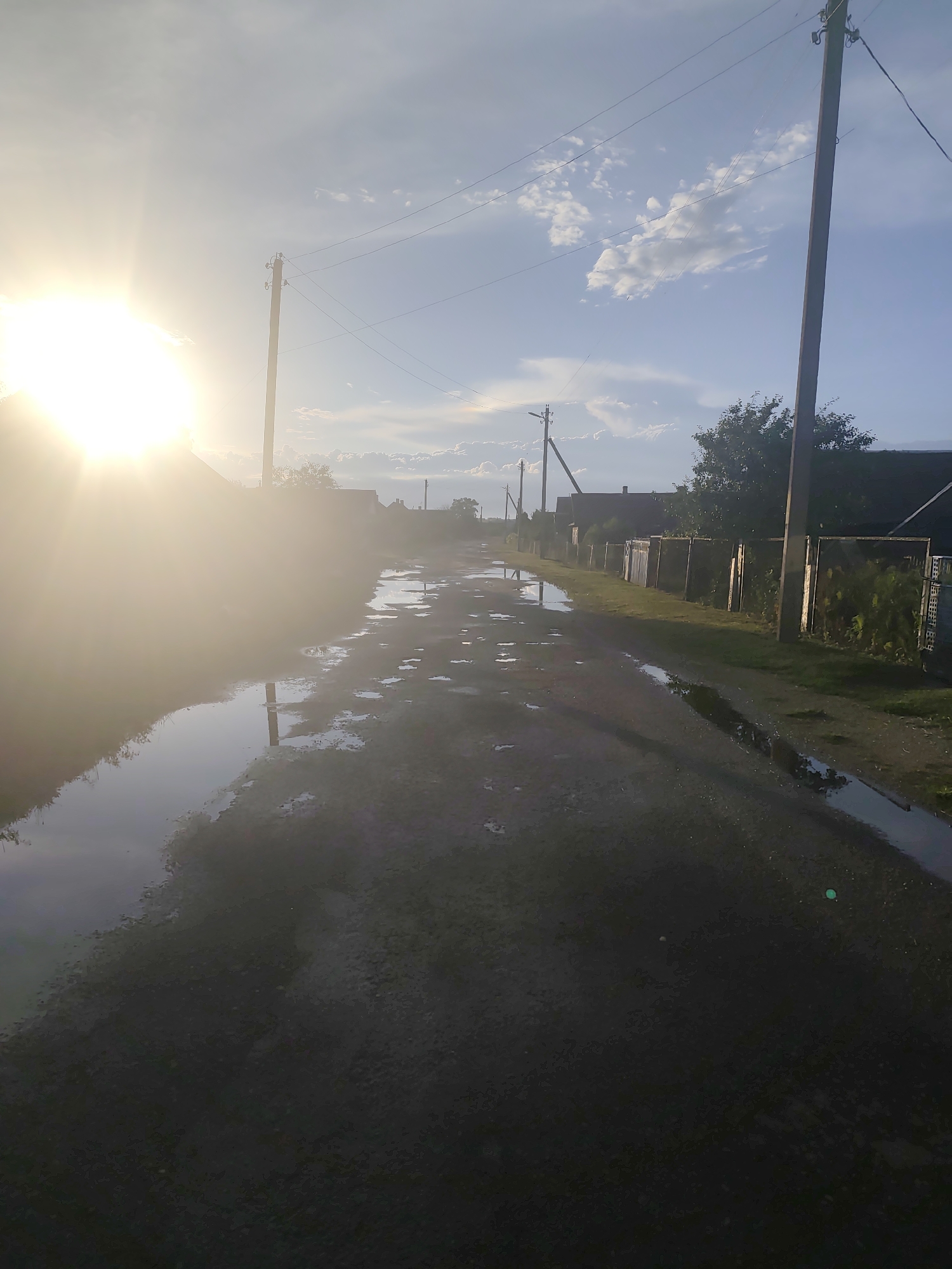 The image depicts a rural road with numerous puddles reflecting the sky and surrounding environment. The sun is prominently visible on the left side, casting a strong glare and creating lens flare. Power lines and utility poles are present, indicating infrastructure in the area. On the right side of the road, there are fences and buildings, suggesting a residential area. The scene appears to be set in the Яглевіцкі сельскі Савет, Belarus, given the context provided. The overall impression is that of a village road after rain, bathed in the warm light of the sun.
