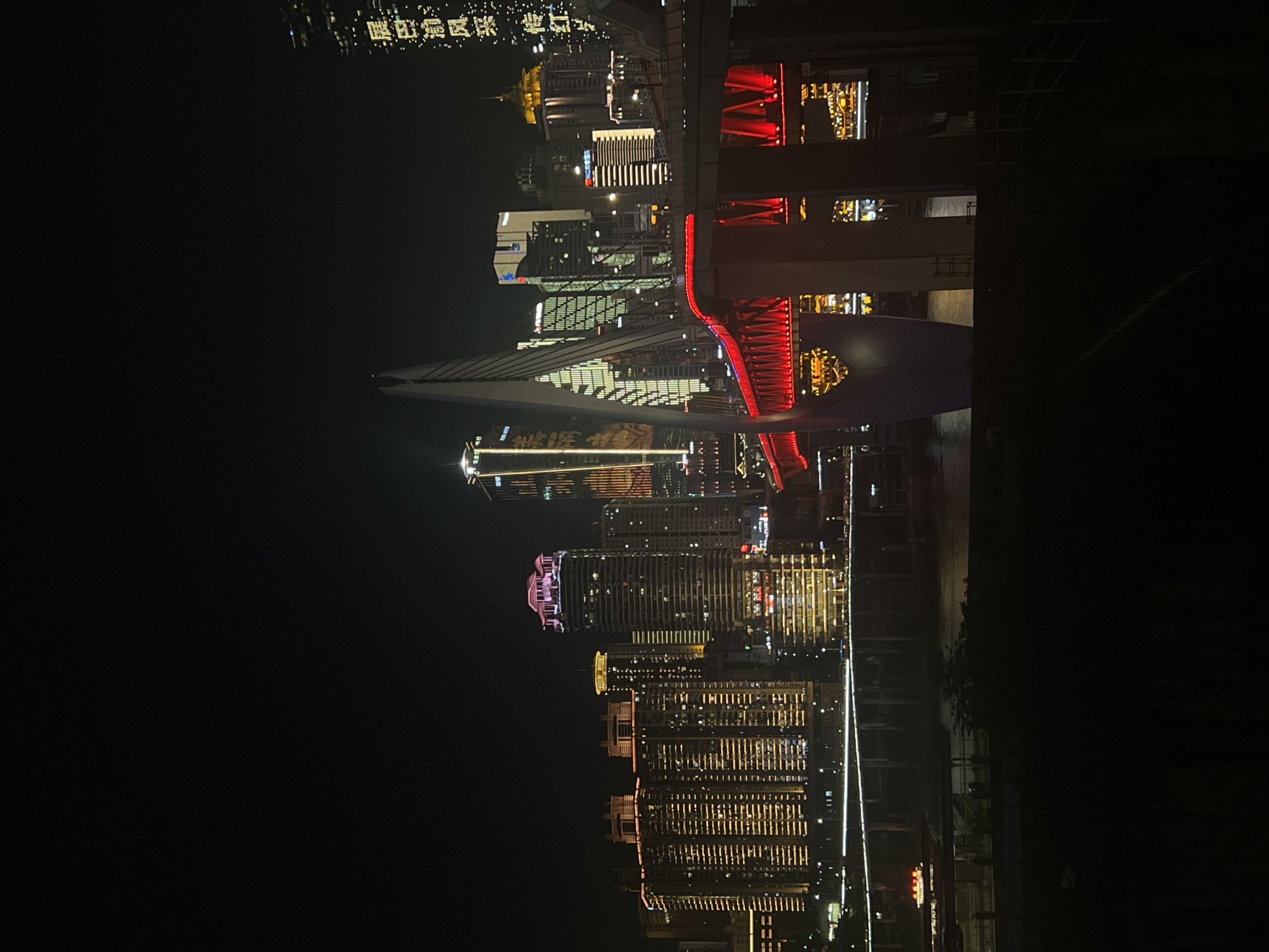 The image captures a nighttime cityscape in Jiangbei Cheng Street, China, dominated by illuminated skyscrapers and a striking red-lit bridge. The foreground is dark, obscuring much of the lower landscape. A multi-level elevated highway, lined with white lights, crosses the midground. To the left of the highway, a dark river reflects some of the city lights, and a small boat with red and white lights is visible.
The central focus is a modern cable-stayed bridge, its arch illuminated with vibrant red lights, creating a dramatic contrast against the dark sky. A large, white, angled pillar, part of the bridge's support structure, rises prominently.
Behind the bridge, a cluster of tall buildings forms the city skyline. Many of these skyscrapers are illuminated with a warm, yellowish light, outlining their windows and architectural features. One building on the far right displays vertical lines of light and vertical Chinese characters in white. Another prominent building to the left of the bridge has a distinctive pink illuminated rooftop. The overall impression is a bustling, modern urban environment at night, with the architecture dramatically lit.
