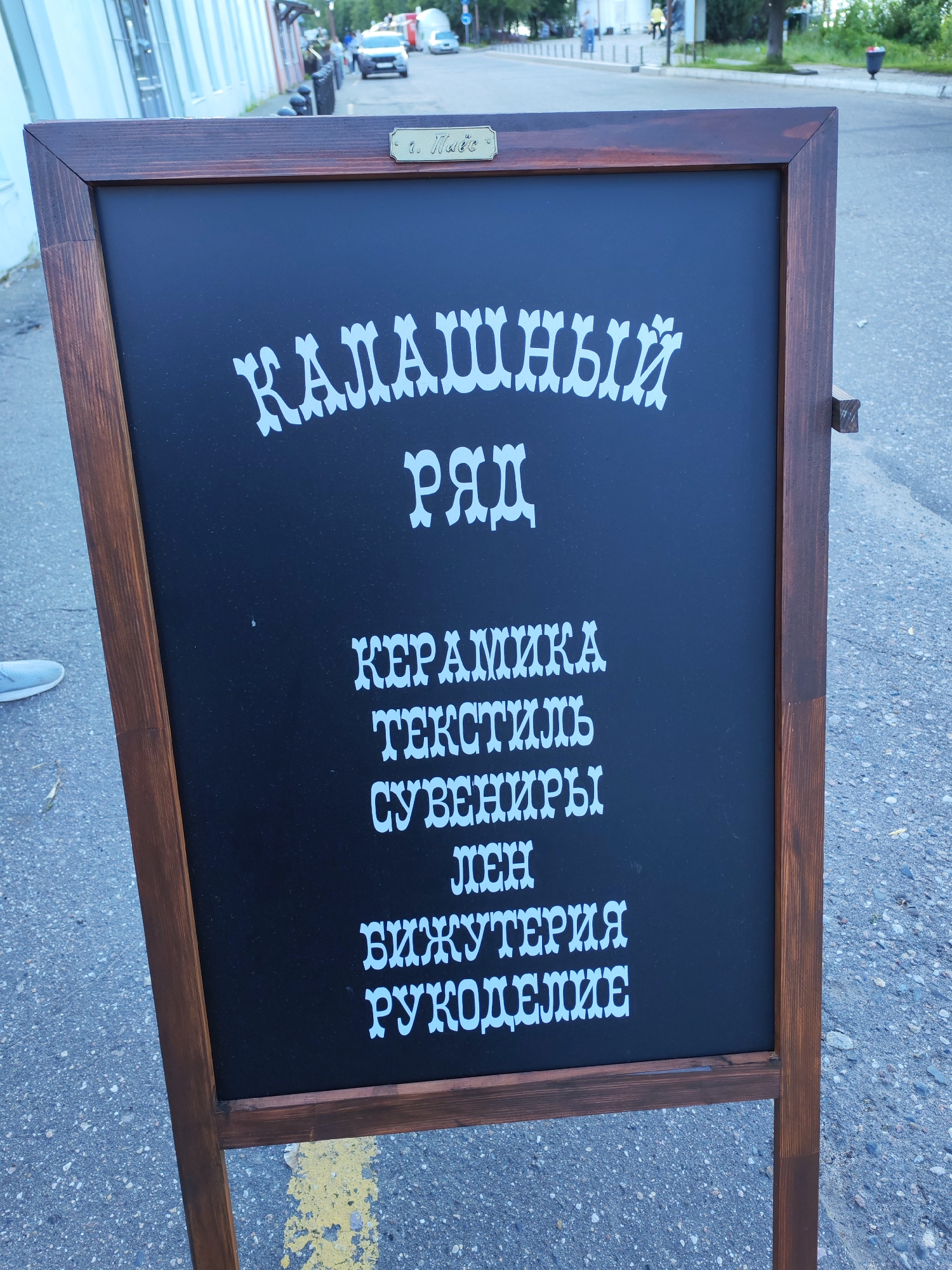 The image shows a black chalkboard sign with white text, placed outdoors on a paved surface with a yellow line painted on it. The sign is framed in dark wood. At the top of the chalkboard, there is a small brass plaque with some engraved text.
The main text on the chalkboard is in a decorative font and appears to be in Russian. It reads:
"КАЛАШНЫЙ
РЯД
КЕРАМИКА
ТЕКСТИЛЬ
СУВЕНИРЫ
ЛЁН
БИЖУТЕРИЯ
РУКОДЕЛИЕ"
This translates to:
"Kalashny
Row
CERAMICS
TEXTILES
SOUVENIRS
LINEN
JEWELRY
HANDICRAFTS"
In the background, a street scene is visible with buildings on the left and trees and more buildings further down the street. There are cars and a few people walking in the distance. The lighting suggests it is daytime, possibly late afternoon, with overcast or diffused sunlight. The setting appears to be an urban or semi-urban environment, consistent with the provided location context of Plyos, Russia. The overall impression is of a market or a shop advertising a variety of handcrafted and textile goods.