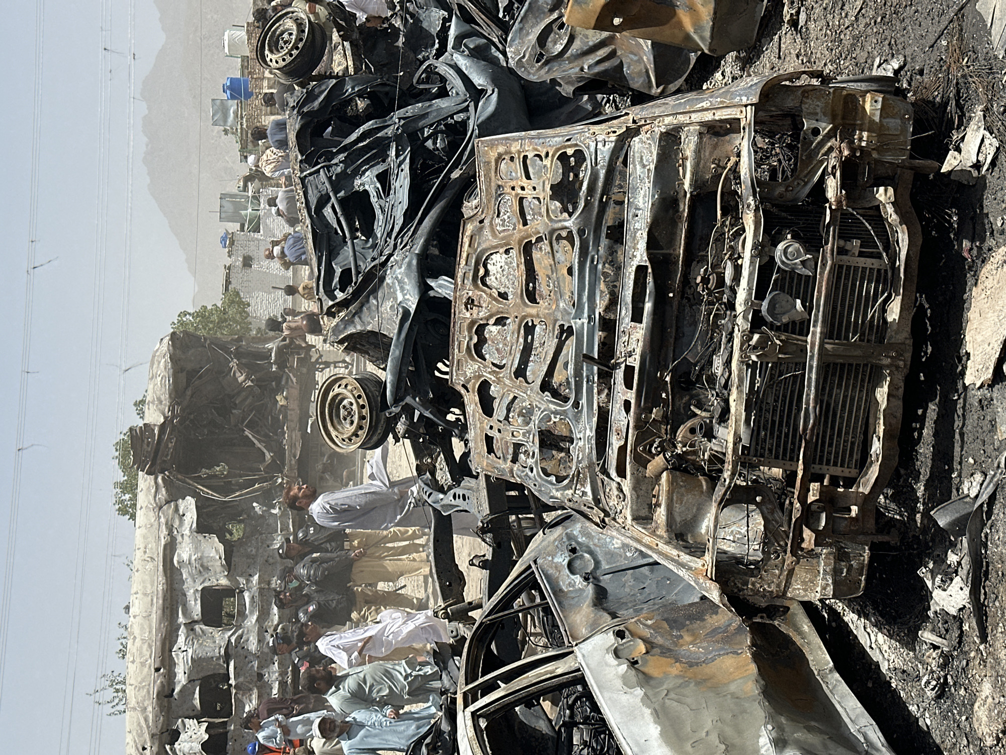The image depicts the aftermath of a catastrophic event involving multiple vehicles, likely a bombing or fire, in Quetta City Tehsil, Pakistan. The scene is dominated by the charred remains of what appear to be cars and a bus. The vehicles are severely damaged, with twisted metal frames, burned interiors, and missing or disintegrated components. A crowd of mostly men, many wearing traditional shalwar kameez, stand nearby, observing the wreckage. Some individuals are dressed in what appears to be police uniforms. The background shows buildings and power lines, suggesting an urban or semi-urban location, with distant mountains visible through a hazy atmosphere. The environment is dusty and the air appears thick, possibly due to smoke or debris.