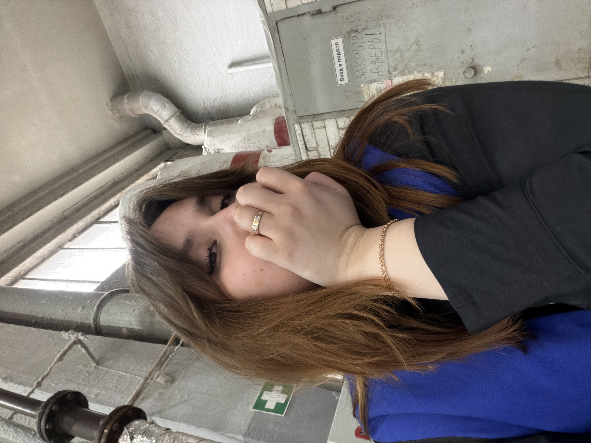 A young woman with long brown hair is posing for a photograph. She is wearing a blue top under a black jacket, with a gold ring on her finger and a gold bracelet on her wrist. The background appears to be an industrial or utility space. Visible elements include pipes, a window, and a gray utility box with a partially legible Russian inscription that reads "ЗАКРЫВАЙ ДВЕРИ!". There is also a green and white first aid symbol visible on a surface below the woman. Given the inscription is in Russian, it is likely that this photograph was taken somewhere in Russia. Based on the general location of photographs on this user's profile, the image was likely taken in Chaikovsky, Russia.