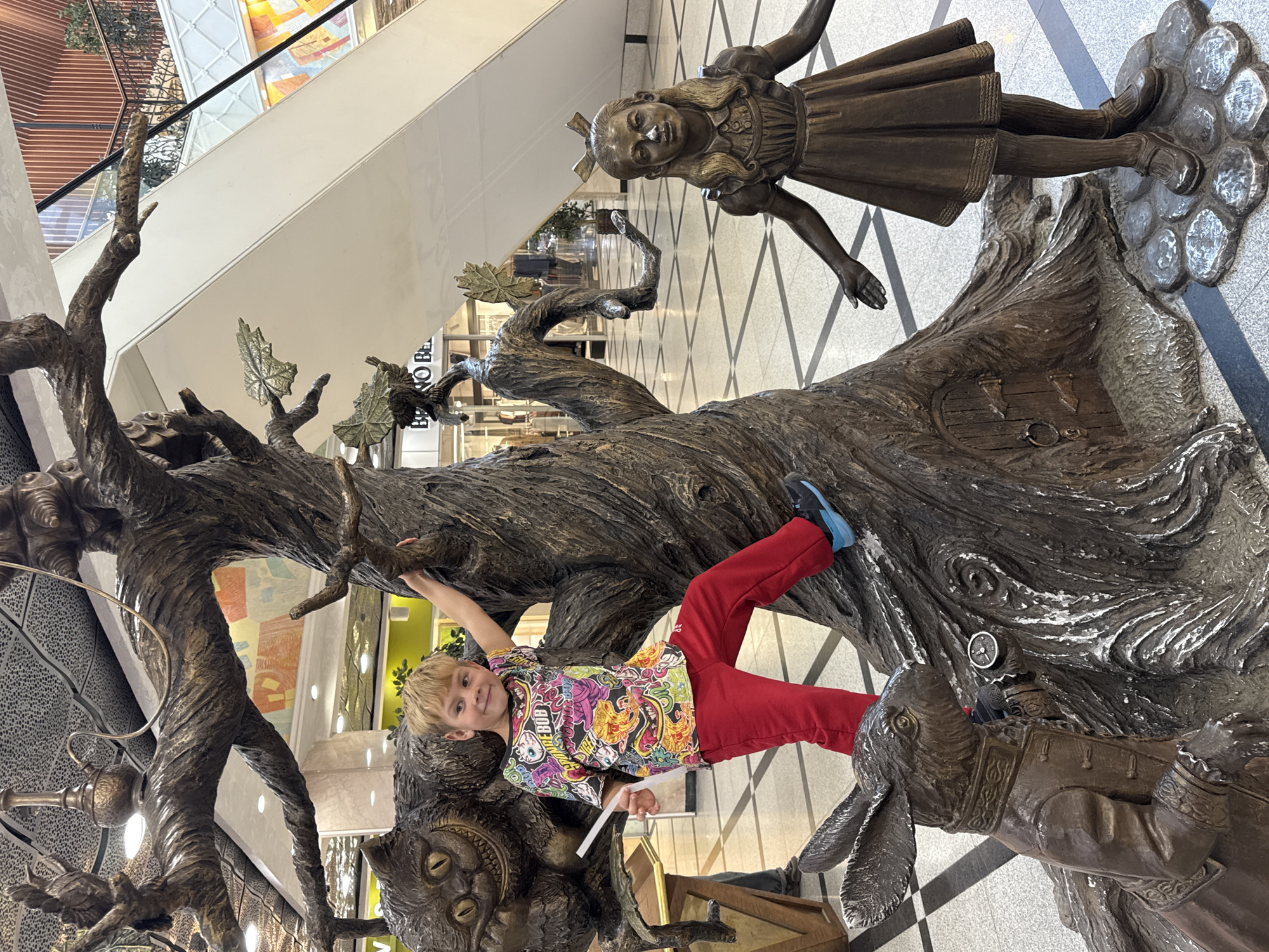 A boy in red pants and a colorful shirt is climbing on a large, bronze-colored sculpture of characters from "Alice in Wonderland." The sculpture includes a large tree, Alice, the Cheshire Cat, and the White Rabbit. The sculpture is indoors in what appears to be a shopping mall, as evidenced by the escalators and retail storefronts in the background. The location is Yekaterinburg, Russia. The boy is smiling and appears to be posing for a photo. The sculpture is detailed and includes various textures to resemble wood, fur, and clothing.