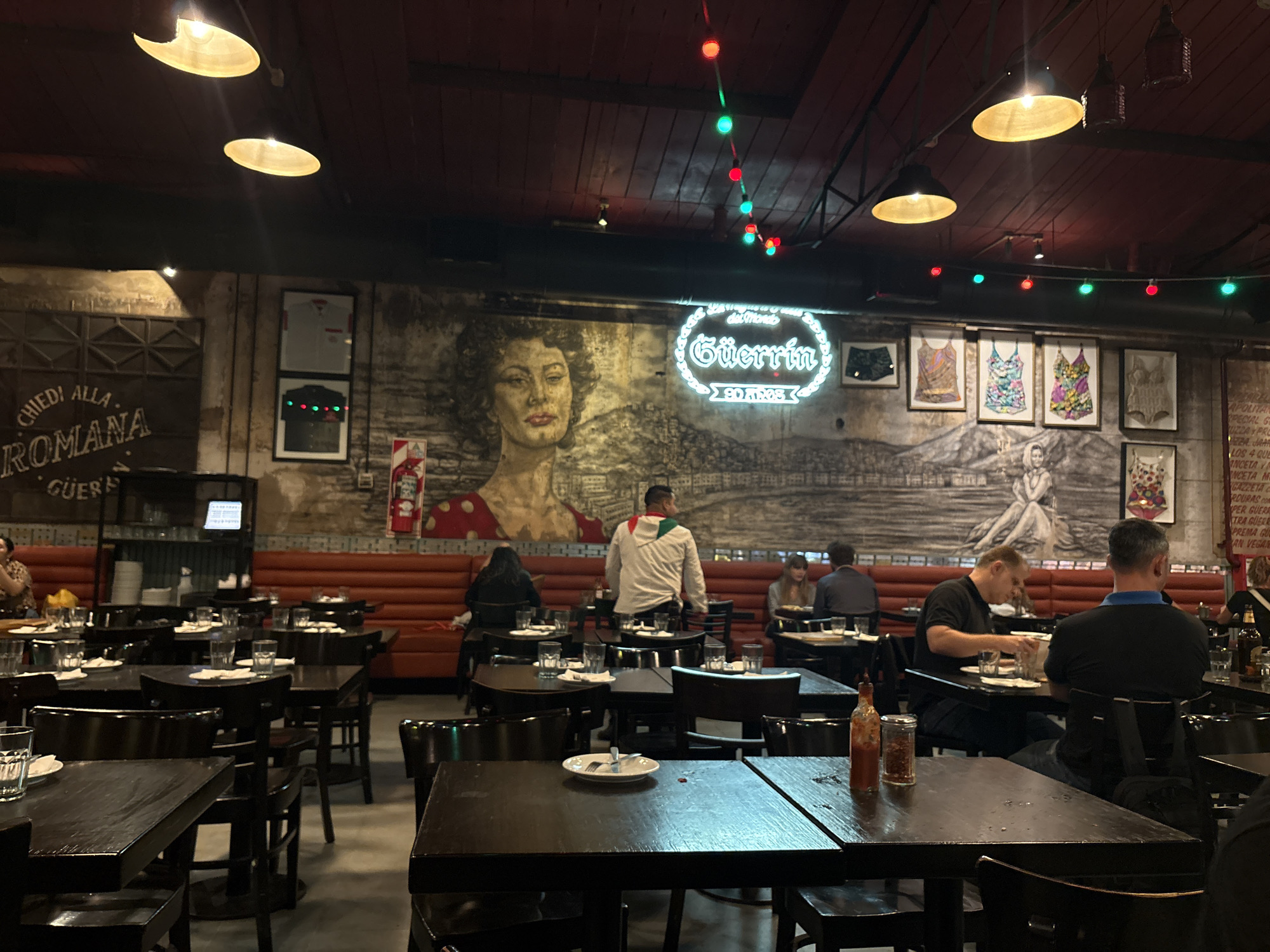 The image depicts the interior of Pizzería Güerrín, a restaurant located on Corrientes Avenue in Buenos Aires, Argentina. The scene is an active, artificially lit dining area.
The background features a textured wall adorned with murals and framed items. On the left, a large mural portrays a woman with dark curly hair, red lipstick, and a red polka-dot garment. Adjacent to this, a panoramic mural depicts a landscape with a figure in the foreground. A prominent white neon sign in script font spells "Güerrín," with "90 AÑOS" visible below it. Other wall decor includes framed shirts (one white, one dark) on the left, and multiple framed colorful swimsuits or bodysuits with floral patterns on the right. A red fire extinguisher is mounted on the far left wall. The dark ceiling is equipped with several overhead pendant lights emitting a warm glow, and a string of red and green decorative lights. Along the back wall, a continuous red-orange upholstered bench provides seating.
The foreground and midground are occupied by numerous dark wooden tables with matching chairs. Many tables are set with drinking glasses and white plates; some plates contain cutlery.
Approximately ten to fifteen individuals are visible. Most are seated at tables, engaged in dining or conversation. One individual in a white jacket with red and green stripes is standing in the midground, positioned between tables. On a table in the immediate foreground, a bottle of hot sauce and a glass shaker containing red pepper flakes are visible next to a white plate with cutlery.