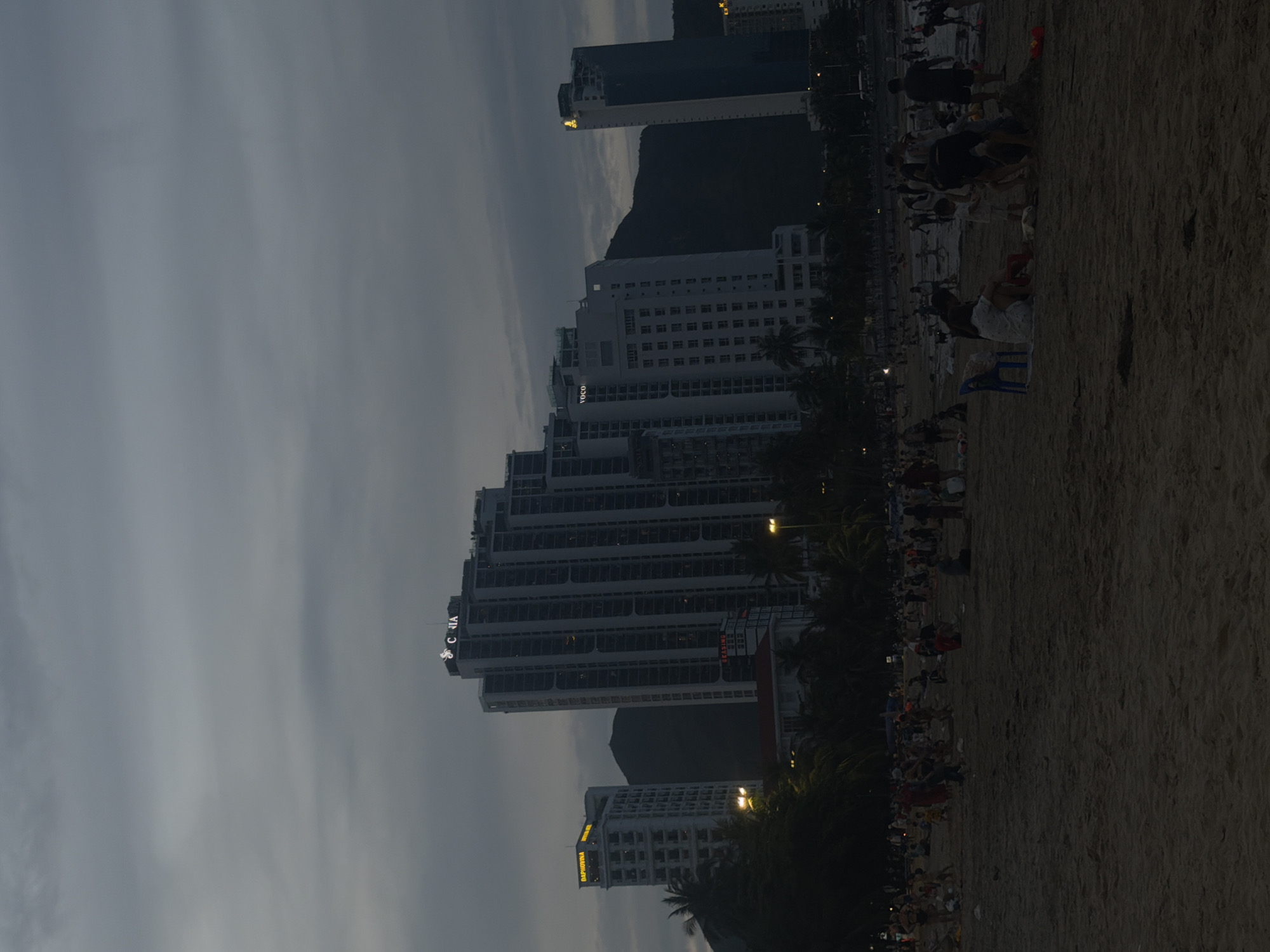 The image depicts a sprawling coastal scene at what appears to be dusk or night, with a sandy beach in the foreground and a cityscape featuring numerous high-rise buildings backed by mountains in the background. The sky is overcast and dark, illuminated faintly.
On the left side of the image, a series of modern high-rise buildings dominate the skyline, suggesting a bustling urban environment along the coast. Many of these structures are lit up, revealing architectural details and signage. Visible text on the buildings includes "VIETRAVEL", "DACAPINOTUNA", "CARIA", and part of "YOC", all illuminated, likely indicating hotels or businesses. Palm trees are scattered between the buildings and along the edge of the beach.
The right side of the image features a wide stretch of sandy beach where numerous people are gathered. Due to the low light conditions, specific activities are hard to discern, but individuals and groups appear to be walking, standing, or sitting on the sand. Some are seen on chairs, while others are simply enjoying the evening atmosphere. The beach appears to be a popular gathering spot.
The overall atmosphere is one of a busy resort town during the evening, transitioning from day to night, with artificial lights beginning to define the urban landscape against the darkening sky and silhouette of the mountains.