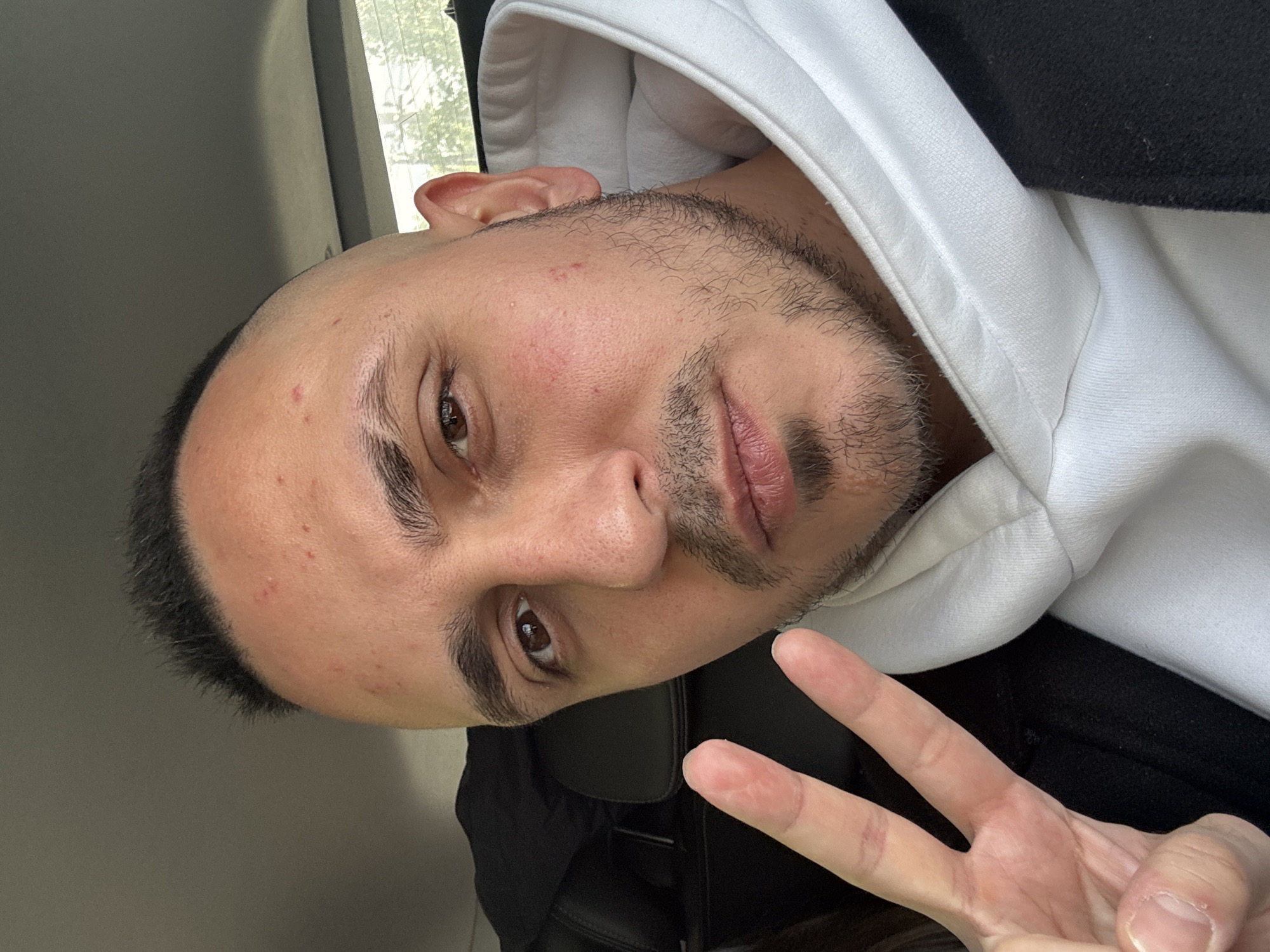 A close-up selfie, rotated counter-clockwise, of a man with light-brown skin and short, dark hair. He has a trimmed beard and mustache and brown eyes. He is wearing a white hoodie and a black jacket. His left hand is visible, making a peace sign. The background suggests the interior of a car, possibly in Pargolovo, Russia, though no specific landmarks are discernible. The car is located outdoors during daylight, possibly near some trees outside the car.