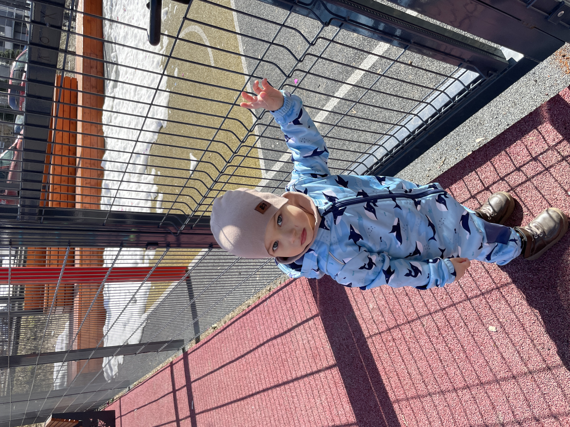 A child in a blue and black whale-patterned snowsuit and a beige hat stands on a red, textured ground, likely a playground surface. The child is near a black metal fence, reaching a hand toward the fence. Behind the fence, there is snow and a wooden structure. The location is Izhevsk, Russia.