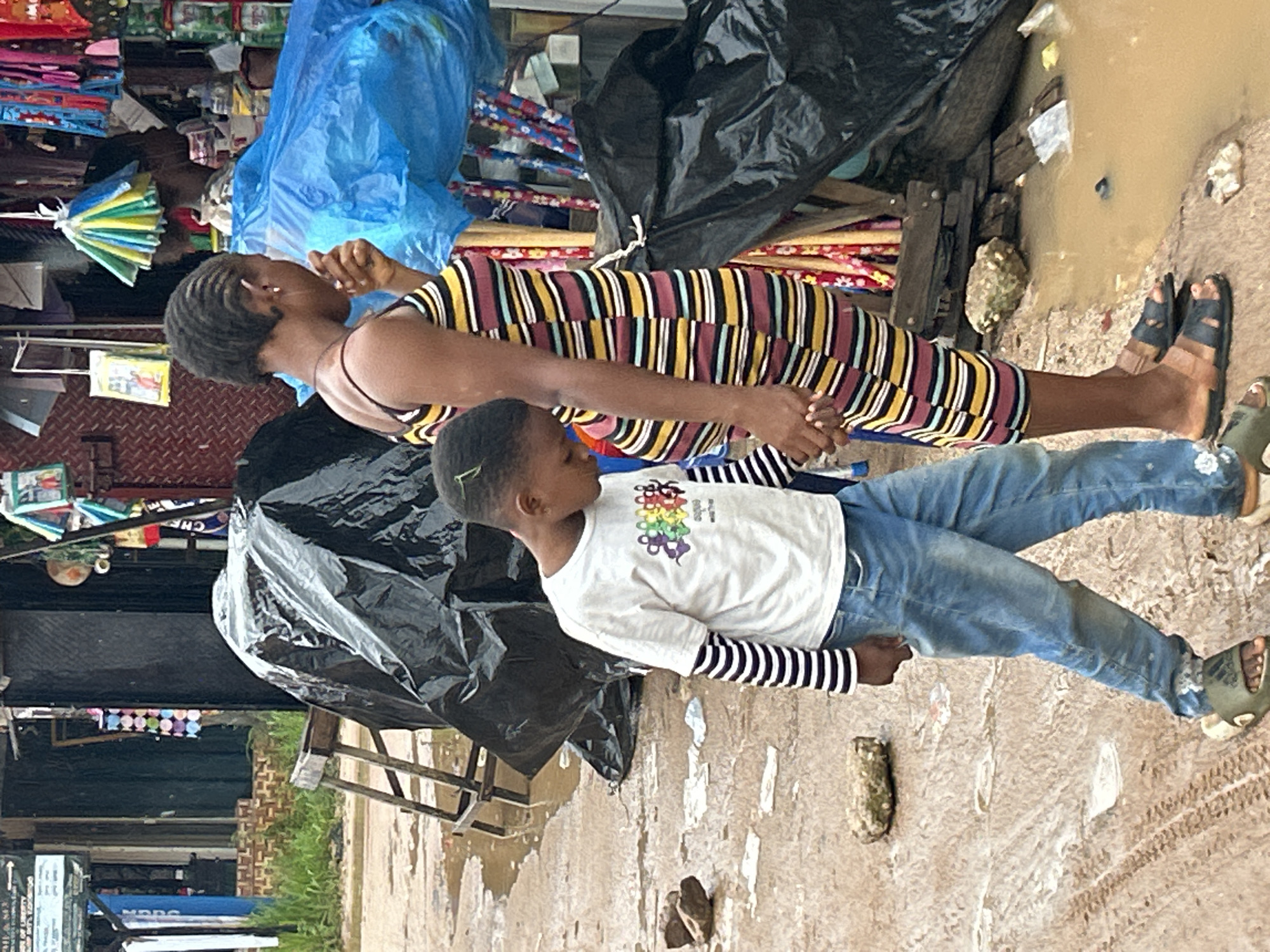 The image depicts an outdoor street or market scene in Ihiagwa, Nigeria, characterized by muddy and wet ground with visible tire tracks and puddles. In the foreground, a dark-skinned woman and a dark-skinned child walk together, holding hands. The woman, positioned on the right, faces left in profile, displaying short braided hair and wearing small hoop earrings. She is dressed in a sleeveless, form-fitting dress with horizontal stripes in maroon, yellow, white, light blue, and dark blue, and appears to be pregnant. Her right hand is raised towards her mouth. She wears dark-colored slide sandals. The child, on the left, looks downward and wears a white short-sleeved t-shirt with a colorful "SQUARE" graphic design over a long-sleeved black and white striped shirt, light blue denim jeans, and olive green Adidas slide sandals. A few green strands are visible in the child's hair.
The background features various market elements. To the left, a partial view of a structure with a blue sign, green vegetation, and stacked bricks is visible. A wooden A-frame structure is present mid-ground, partially obscured by a large black plastic tarp covering an unseen object. To the right, a market stall or shop displays numerous items, including colorful hanging paper umbrellas (yellow, blue, green), various packaged goods, and other merchandise. A bright blue plastic tarp covers part of a display, and additional black plastic tarps are used to cover items closer to the ground on the right. Significant puddles of muddy brown water are visible in the foreground, particularly on the bottom right. Scattered rocks and light-colored debris are present on the ground.