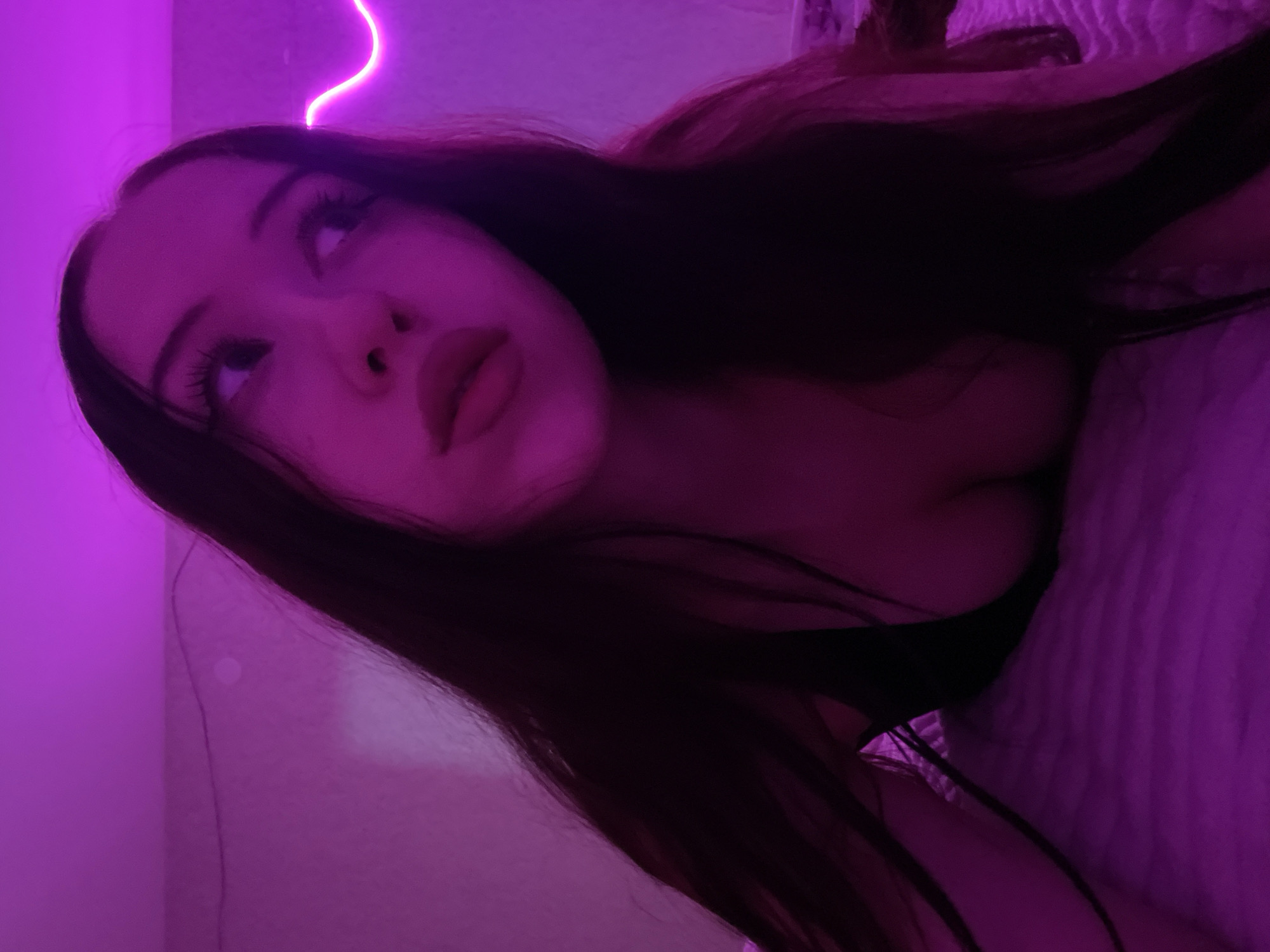 The image shows a young woman lying on her side on a bed. She has long, dark hair and is wearing a dark-colored top. The room is illuminated by purple lighting, with a neon light strip visible in the background. The woman is looking away from the camera with her mouth slightly open. The bed appears to have a textured surface. There are no other people or objects of particular note in the visible area of the image. The location of Votkinsk, Russia, is not depicted or suggested by any elements within the image.