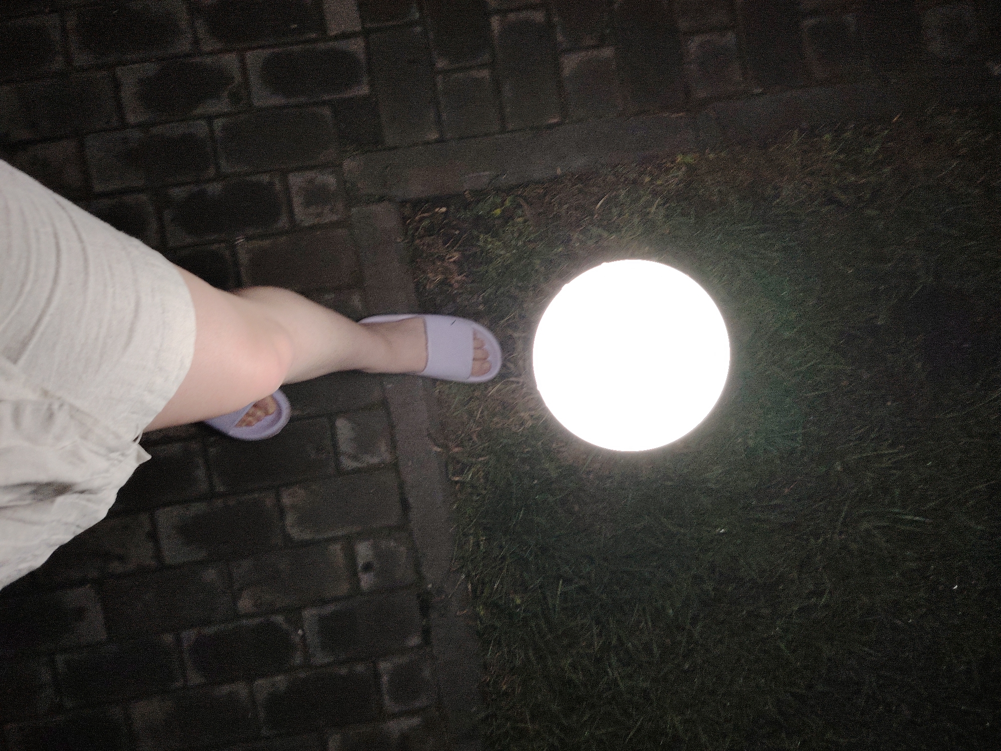 The image presents a nighttime scene outdoors. The ground is paved with dark, possibly wet, rectangular bricks on the left and covered with dark grass on the right. A circular, brightly lit light fixture is positioned on the grass. A person's leg, clad in light-colored clothing and lavender-colored sandals, is partially visible near the brick path and light fixture. No specific landmarks are recognizable that would indicate a precise location within Malyi Bukor, Russia.