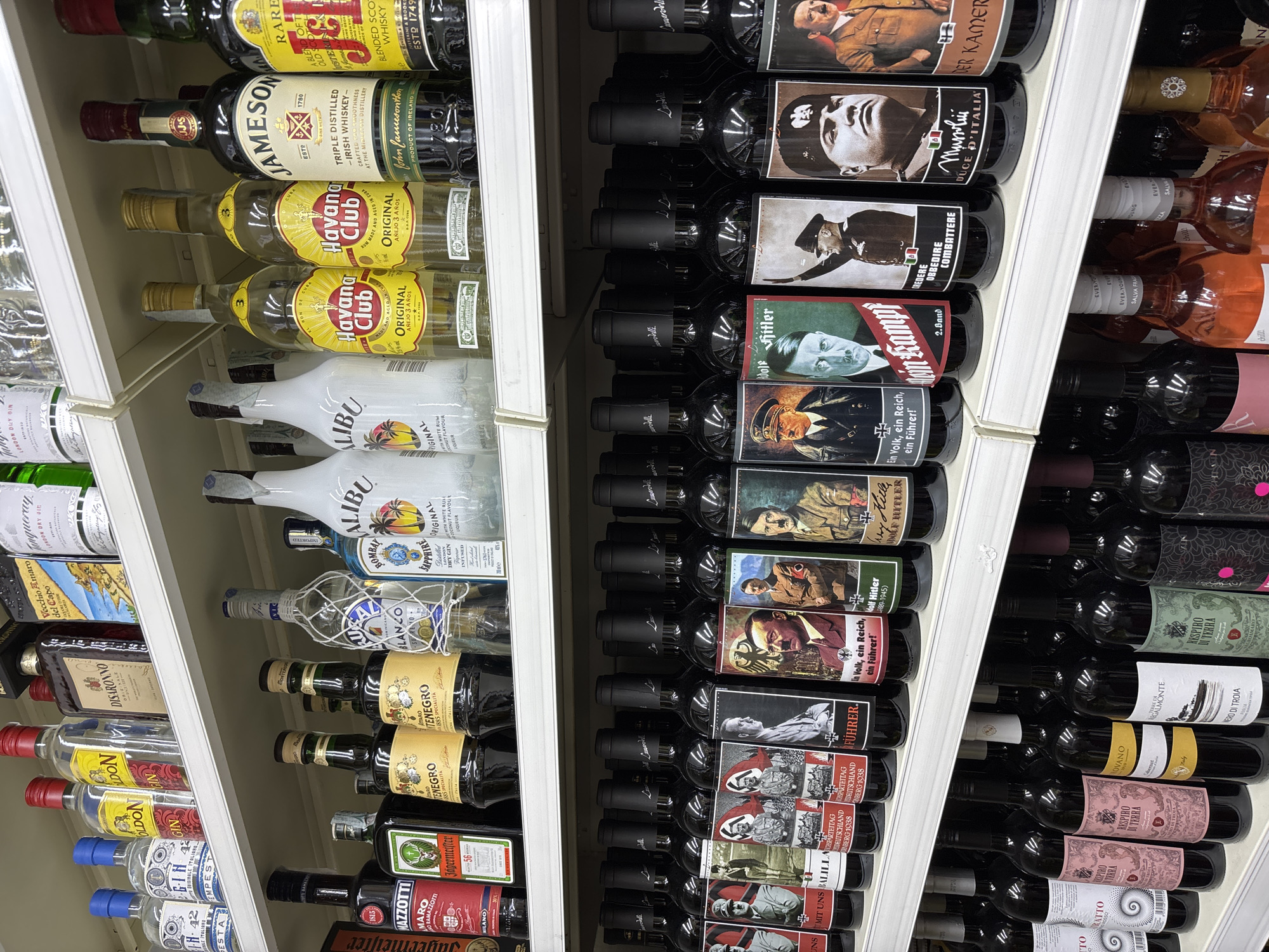 The image depicts multiple shelves stocked with various alcoholic beverages, likely inside a retail store. The left shelves primarily feature bottles of whiskey, rum, gin, and other liquors, including brands like Jameson, Havana Club, Malibu, Tanqueray, and Jägermeister. The right shelves predominantly showcase wine bottles with controversial labels featuring images of Adolf Hitler and Benito Mussolini. It appears this location is in Jesolo, Italy, based on the name on the wine bottle "Mussolini, DUCE D'ITALIA".