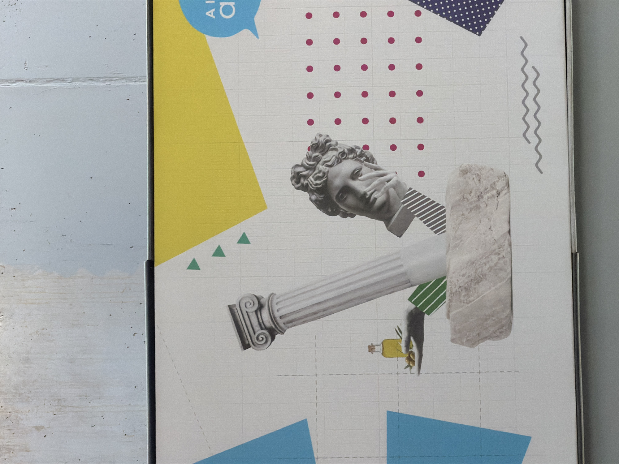 This image shows a poster on a wall next to a gray brick wall. The poster is a collage consisting of a head of a statue in the upper center of the poster and a column protruding horizontally from under the head. The collage also contains a yellow triangle in the upper left, a blue triangle on the bottom, and geometric shapes. A hand is holding a bottle of yellow liquid. The poster features a white grid-like background. The location is likely in Spata-Loutsa Municipal Unit, Greece.