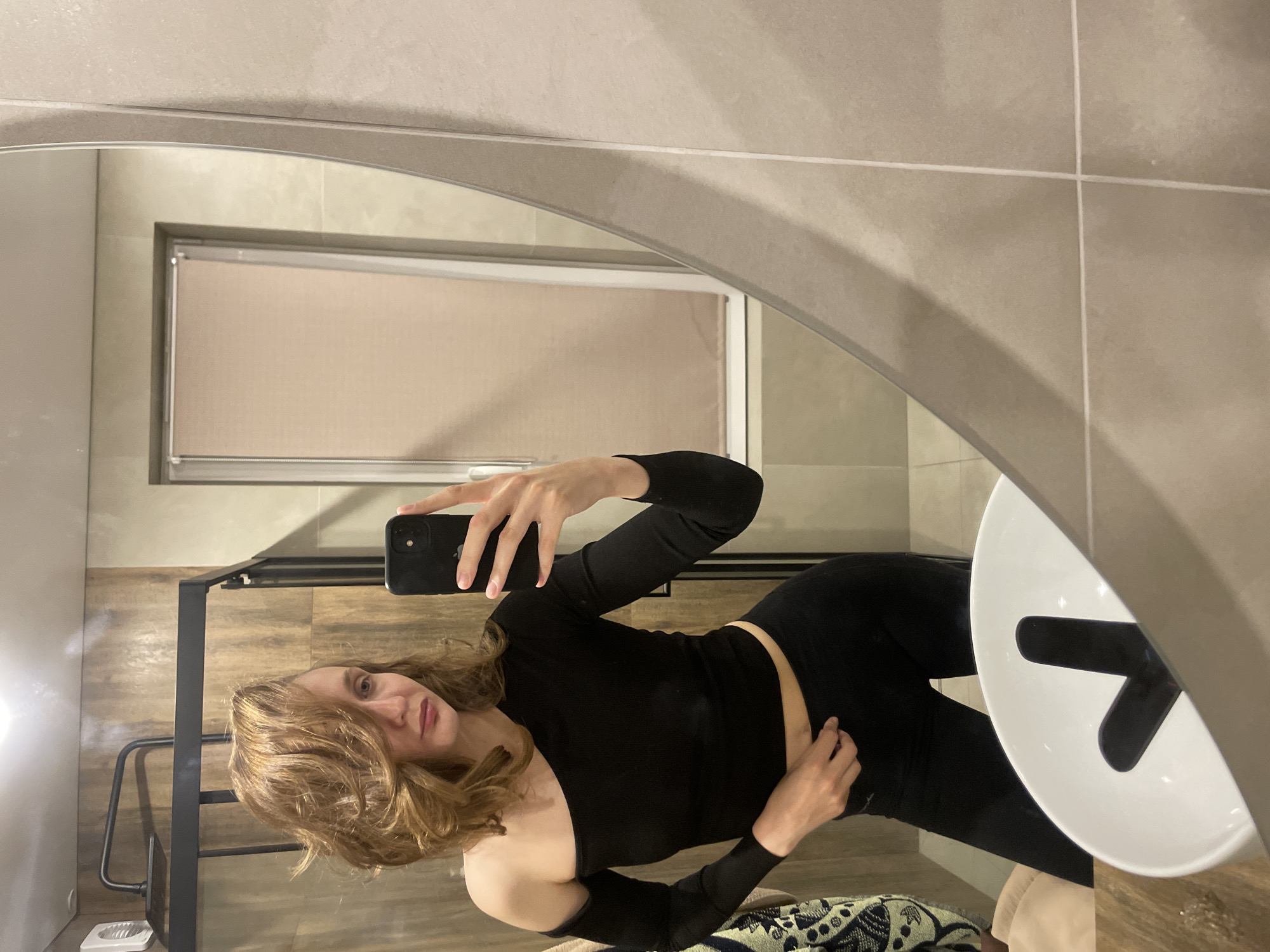 A woman with reddish-brown, wavy hair is captured in a mirror selfie taken in a modern bathroom. The image shows her reflection, where she is holding a black smartphone with a dual-camera setup in her right hand, positioned to take the photo. She is wearing a fitted black long-sleeved top with one shoulder exposed, and black form-fitting pants or leggings. Her left hand is resting on her hip.
The setting appears to be a contemporary bathroom. The walls are covered in large, light beige or light grey rectangular tiles with thin white grout lines. In the reflection, a window with a light-colored blind or shade is visible, suggesting daylight. The floor in the reflection has a wood-like finish.
On the right side of the mirror, a white, modern basin or sink is partially visible, featuring a distinct black cross or T-shaped fixture inside, likely a faucet design. To the left, a portion of a black metal structure, possibly part of a vanity or shelving, is reflected. A small white ceramic object, possibly a bidet or floor drain, is also visible on the floor in the lower left of the reflection. A patterned blanket or towel is peeking from the bottom edge of the frame. The woman has a neutral expression, looking directly at her phone's camera. There is no visible text. The location context is Sovetsko-Nikol'skoe Lesnichestvo, Russia.