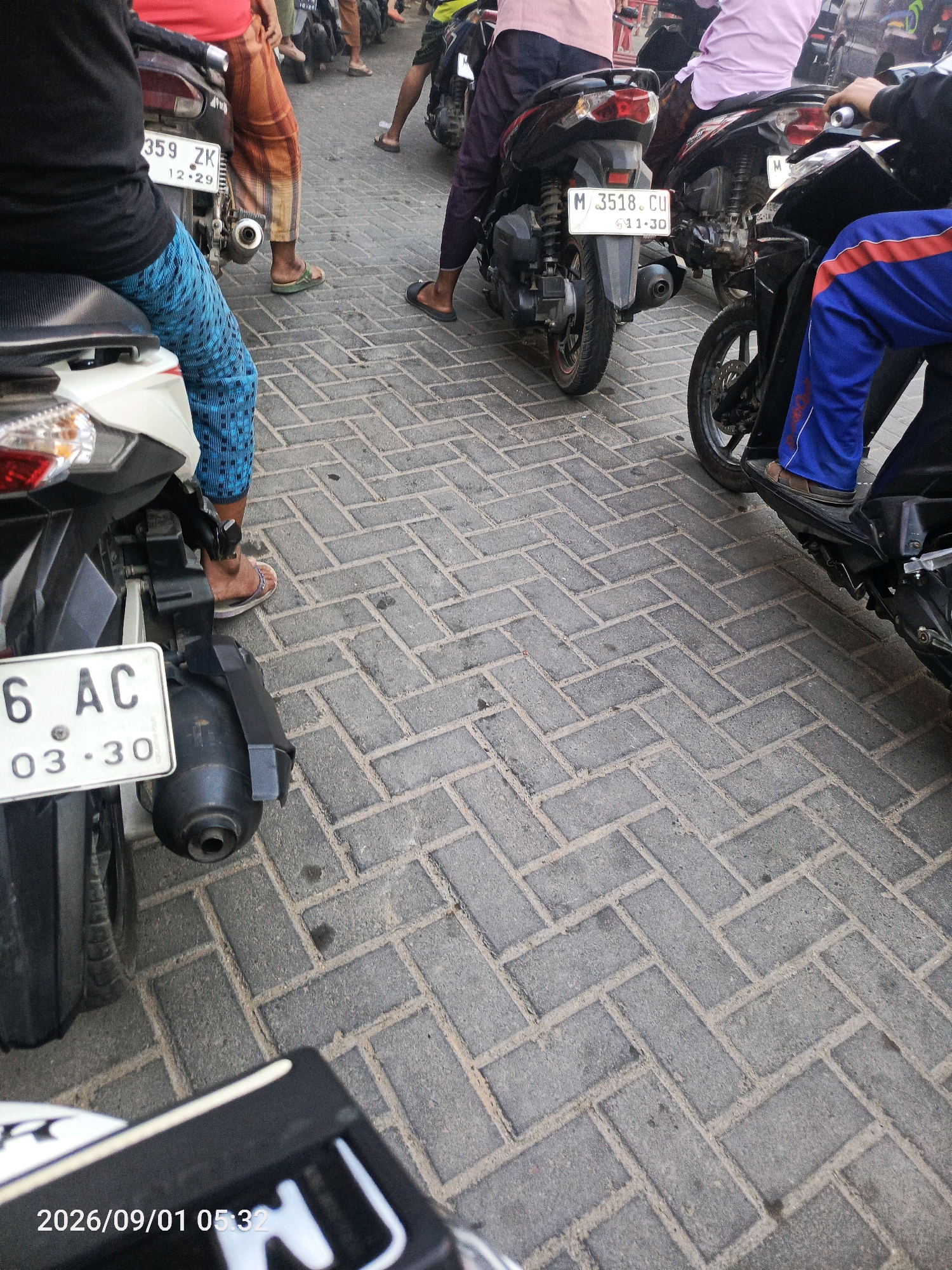 The image shows a close-up view of several motorcycles parked on a brick-patterned paved surface. Two motorcycles are prominently featured in the foreground. The left motorcycle has a white license plate with black text reading "6 AC" and below it "03.30". A portion of a person's legs wearing blue and black pants and flip-flops is visible on the motorcycle seat. The right motorcycle is partially visible with a person wearing blue pants with red stripes and brown sandals. In the background, other motorcycles and individuals are present, with some people seated on their vehicles. The lighting suggests it is daytime. The scene appears to be an outdoor parking area in a city, possibly in Gladak Anyar, Indonesia, given the timestamp at the bottom left corner of the image "2026/09/01 05:32".