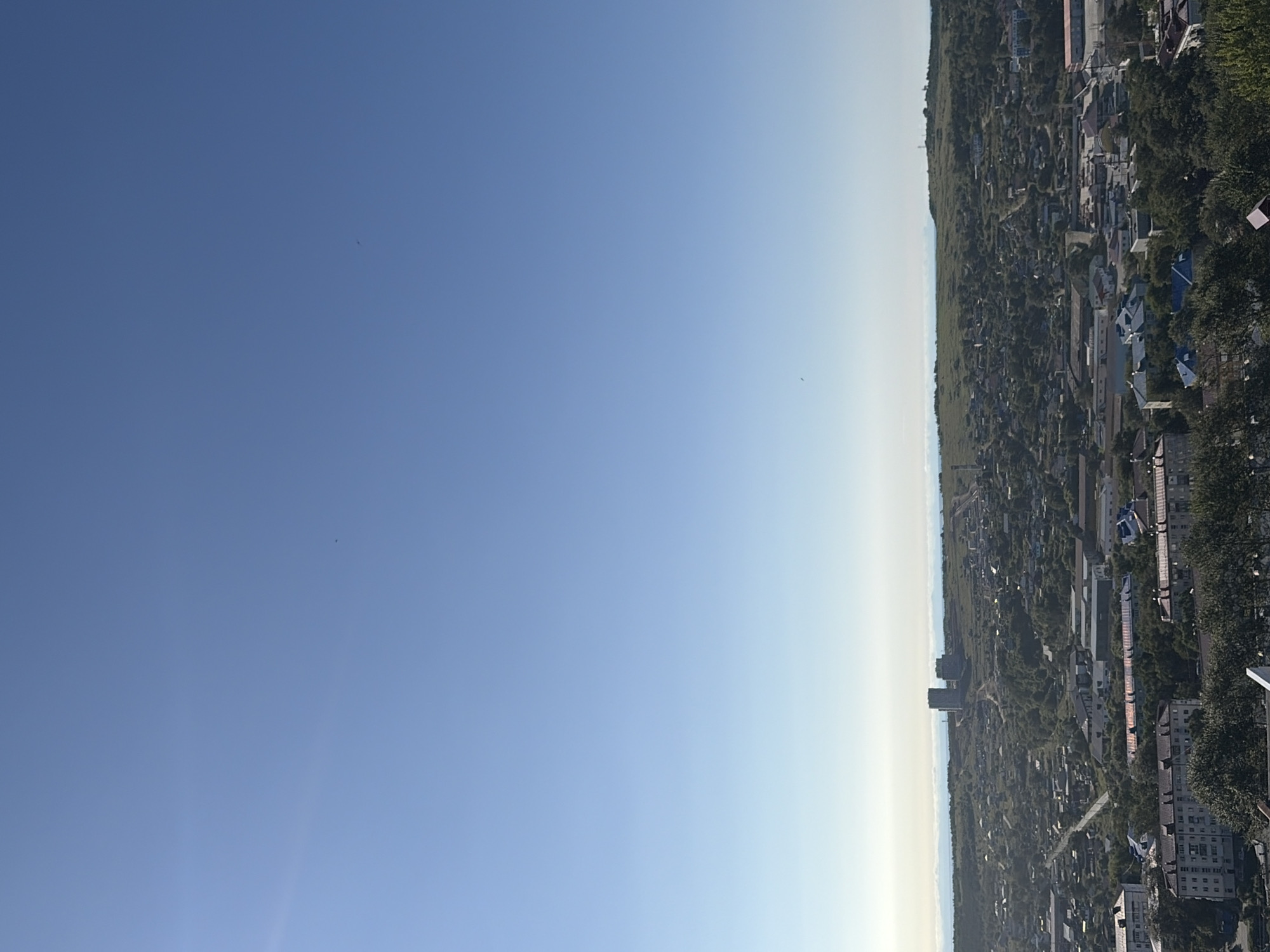 This is an aerial, high-angle shot of a city with a clear blue sky. The city is densely populated with buildings of various sizes, including apartment blocks and industrial structures. Green trees and vegetation are interspersed throughout the urban landscape. In the distance, a body of water is visible, meeting the horizon under a hazy sky. Two tall, distinct buildings stand out on a hill in the background. The overall scene suggests a calm, sunny day in a Russian city.