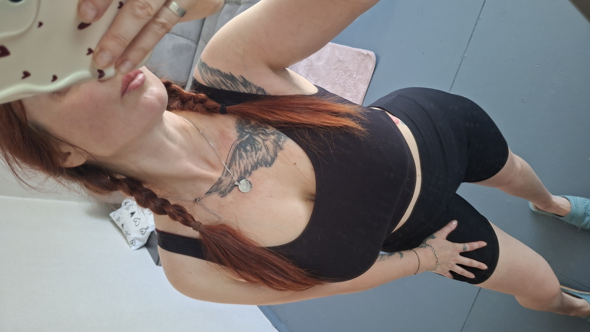 In an indoor setting, a woman with red hair styled in two braids poses for a selfie. She is wearing a black tank top and black biker shorts. A silver necklace with a circular pendant rests on her chest, and a tattoo of wings is visible on her upper chest and shoulder. She is also wearing a silver ring on her right hand and a bracelet on her left wrist. Her left hand is resting on her thigh. In the background, a blurred grey sofa and a pink rug are visible. The floor appears to be tiled or painted blue-grey. The room is sparsely furnished, with a white tissue box with black heart patterns on a surface to the left of the woman. The context of the location in Myadzyel, Belarus, is not visually supported by the image content.