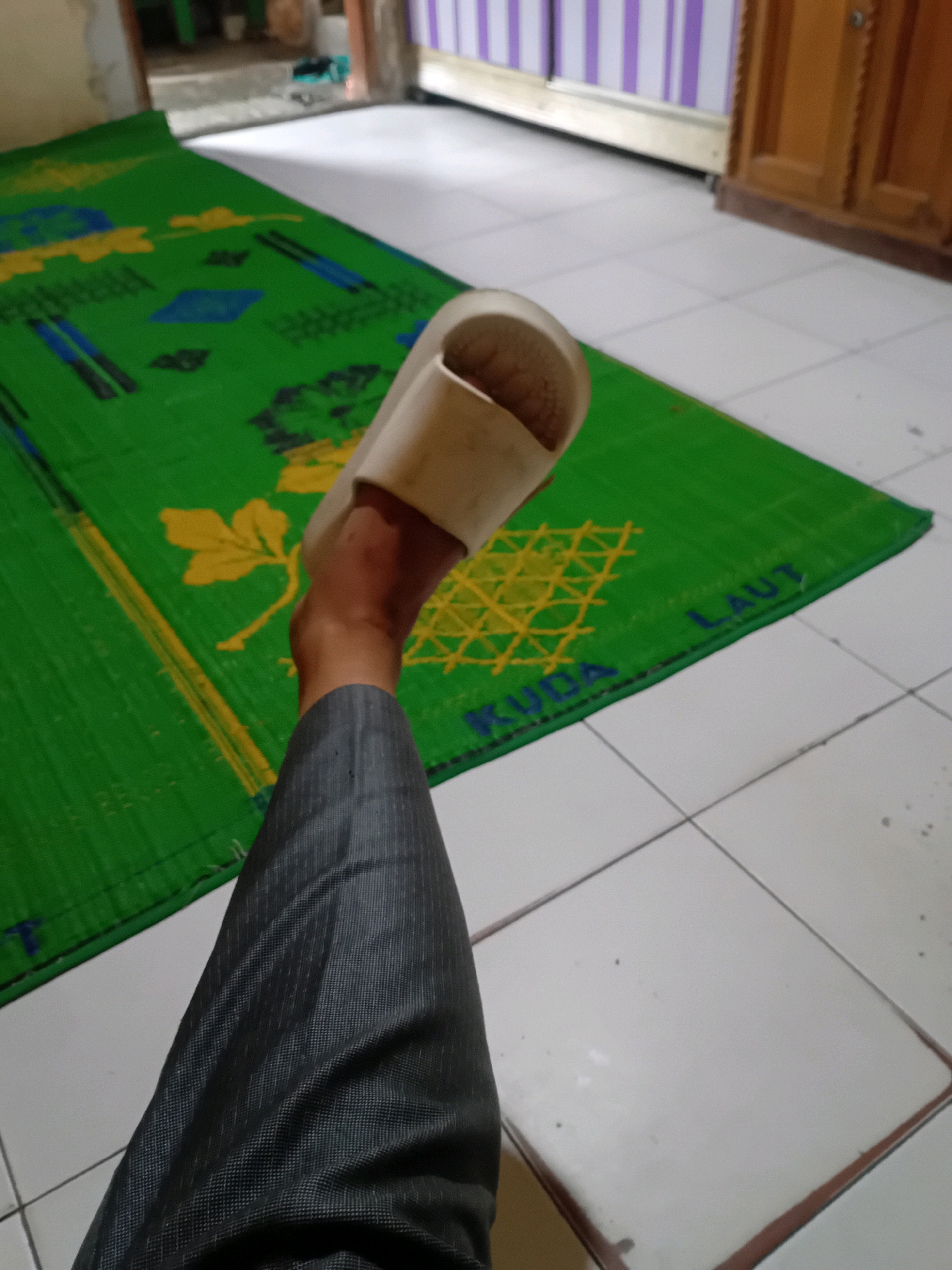 A person's leg, wearing a dark gray pinstripe pant and a white sandal, is positioned on a green mat with yellow and blue patterns. The mat is laid out on a white tiled floor. The leg is bent at the knee, with the foot wearing the sandal resting on the mat. The mat has text printed on it, partially visible as "KUDA LAUT". In the background, there is a wooden cabinet and a wall with purple and white striped decor. The scene appears to be indoors, possibly in a residential setting in Pekanbaru, Indonesia, given the common use of such mats in the region.
