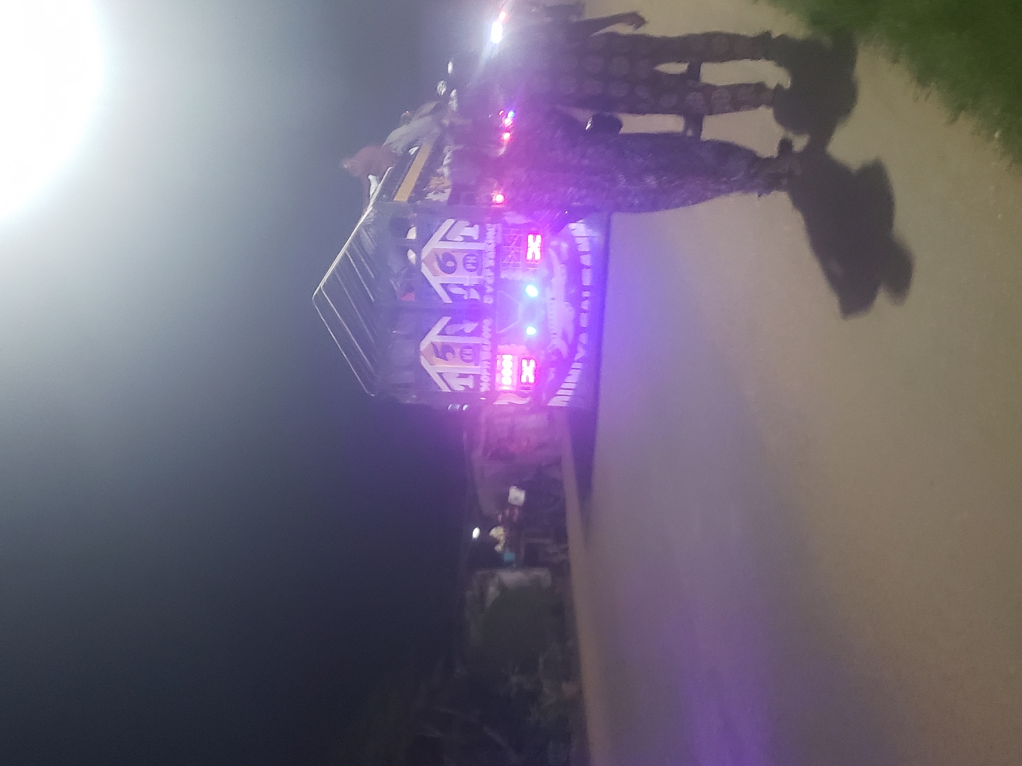 The image depicts a vehicle with illuminated lights in Alalubosa, Nigeria. The vehicle has a rack structure on top. Two individuals are near the vehicle. The scene is at night on a paved surface, possibly a road. Vegetation is visible on the right side of the image.
