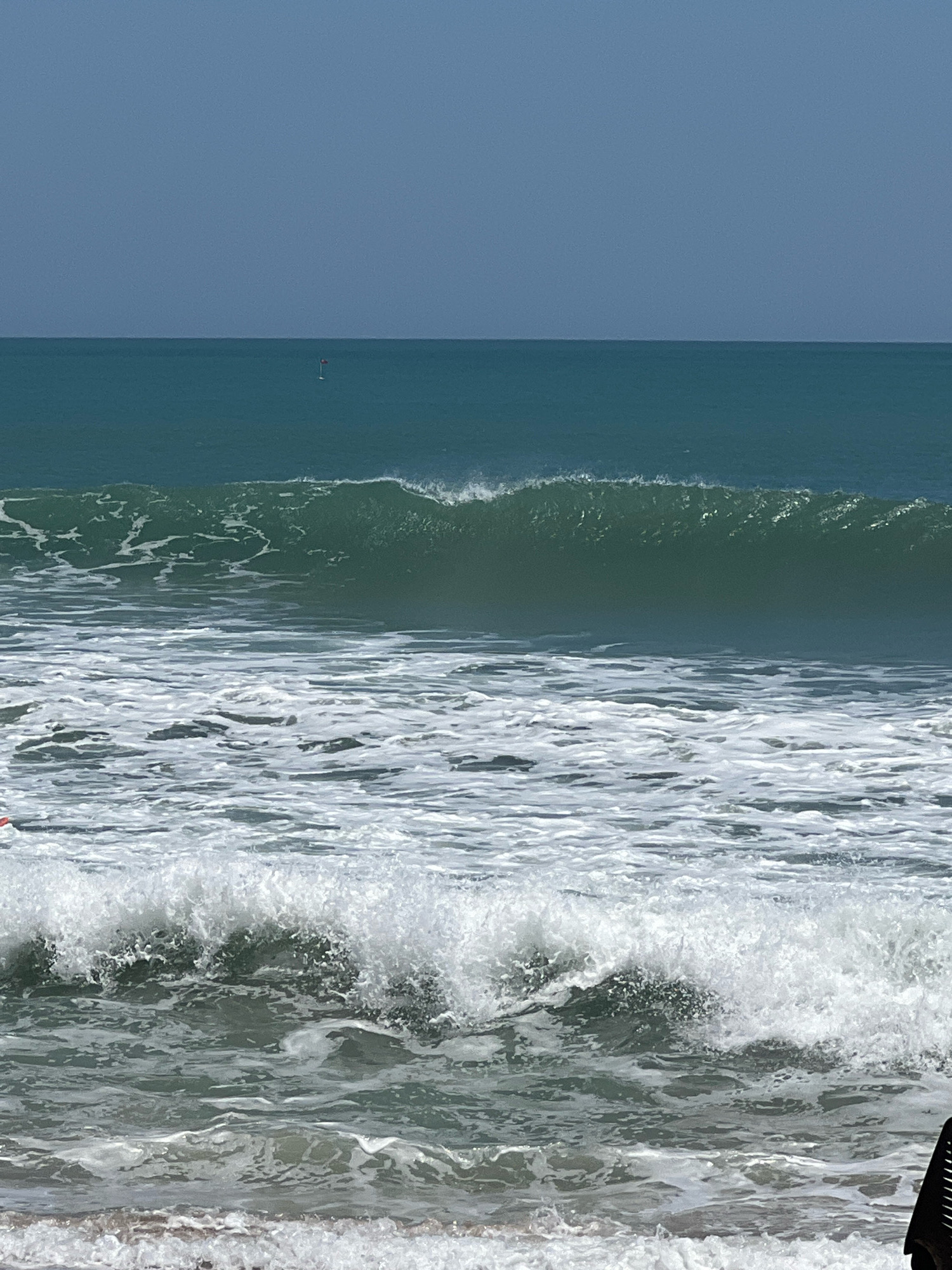 The image captures a scenic view of the ocean in Kuta, Indonesia. The sea is a deep teal with moderate waves, some of which are breaking and forming white foam. The sky above is a clear, pale blue. In the distance, a small red buoy with a white pole is visible. The foreground shows the turbulent surface of the water with whitecaps and foam, indicative of ongoing wave action. Part of a person's orange shirt is visible on the left edge of the frame, and a black object, possibly part of a beach chair or equipment, is visible on the bottom right corner.