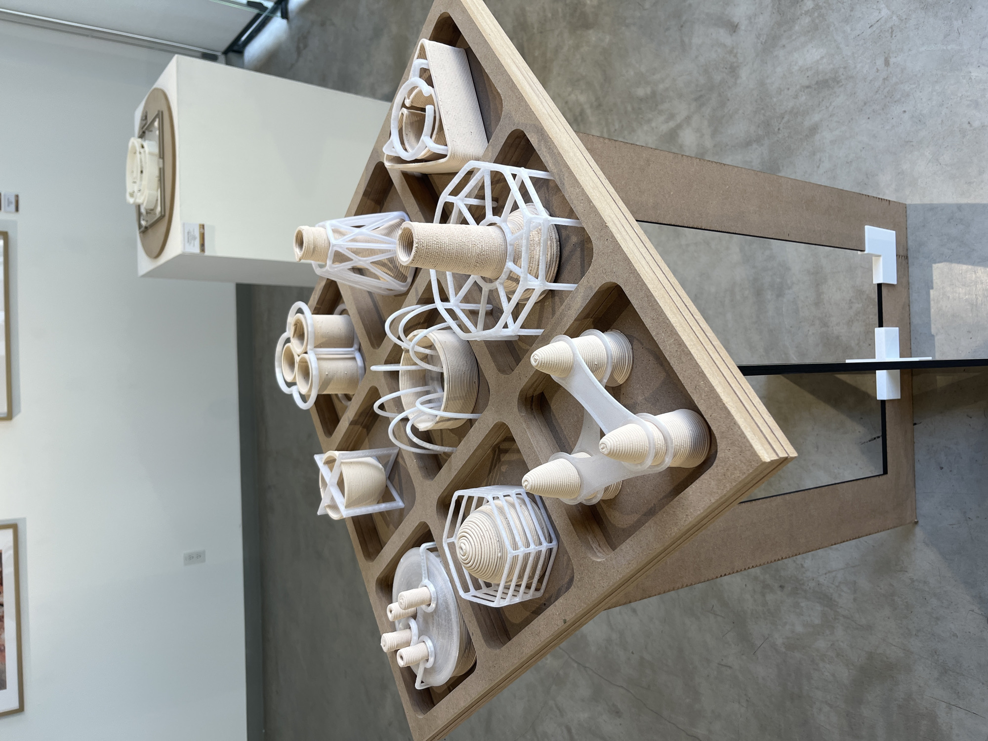 The image depicts an indoor exhibition or gallery space in San Andrés Cholula, Mexico. The central subject is a square display table constructed from light brown wood-composite material (MDF), positioned in the foreground and viewed from a slightly elevated angle. The table's surface features a grid of twelve recessed square compartments, each containing a unique sculptural model.
These models are predominantly light beige or tan with visible layered textures, characteristic of 3D-printed objects. Many incorporate white, translucent, open-framework support structures or cages, which cradle or surround the core beige elements. The models exhibit diverse geometric forms, including conical spirals, tall cylinders with spoked bases, multiple smaller cylinders on flat platforms, and abstract arrangements. For example, one model consists of a conical spiral within a hexagonal white frame, while another presents a tall, textured cylinder secured by a wide, spoked white base.
The display table itself rests on a matching MDF base, which includes vertical panels, reflective mirrored inserts, and white plastic bracket connectors visible at the bottom.
In the background, a white pedestal is positioned to the upper right, supporting another white, possibly 3D-printed, object set on a circular brown base. The walls are plain white, featuring a visible electrical outlet. Partially visible framed artwork hangs on the wall further in the background. The floor is light gray concrete. The lighting is bright and even, consistent with an indoor exhibition environment. No people, actions, or direct interactions are visible in the scene.