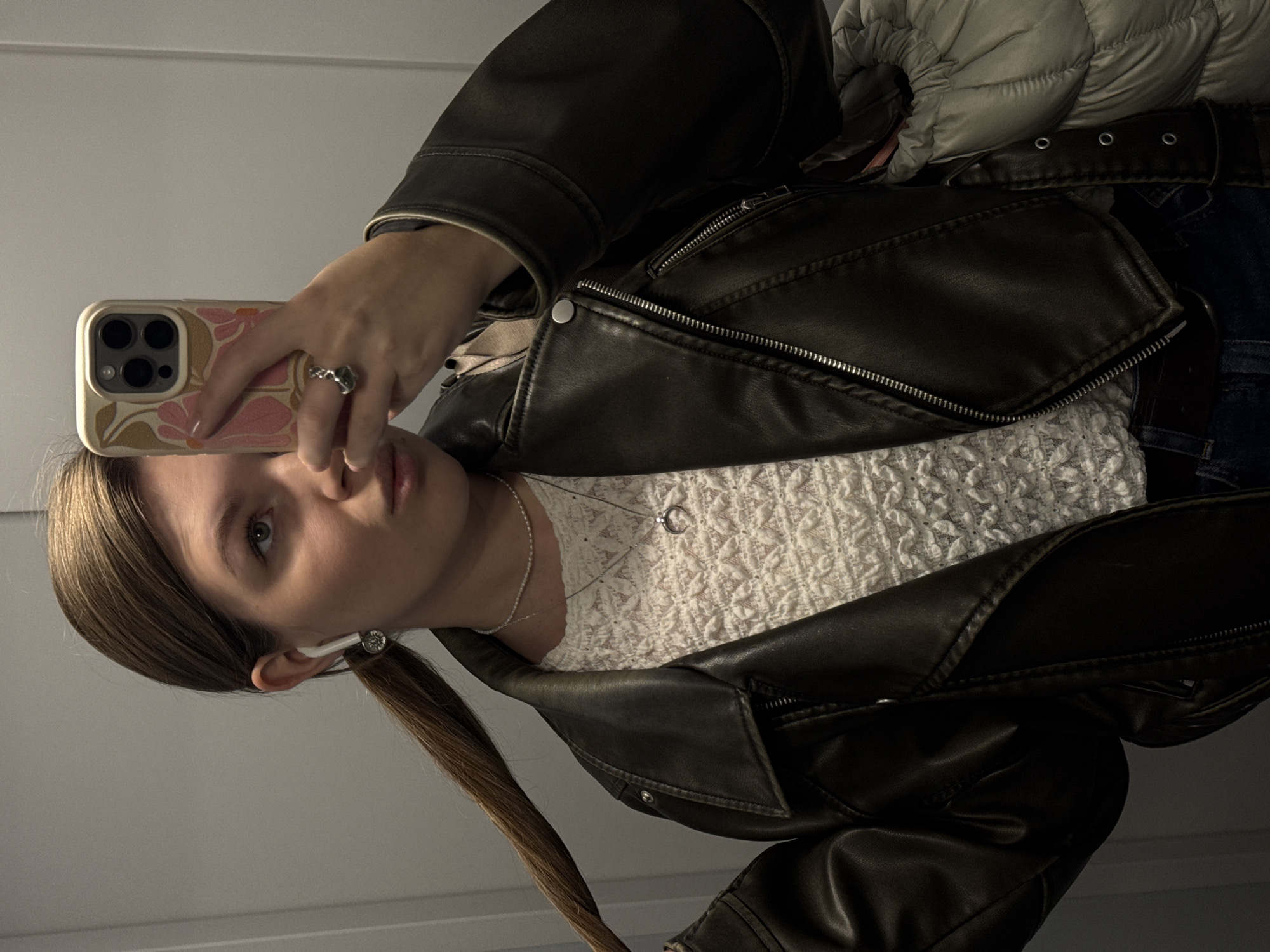 A mirror selfie taken indoors captures a young person from the chest up. The individual, appearing to be female, looks directly into the camera with a neutral expression. She has light-colored eyes and long, light brown hair styled partly in a low ponytail, secured with a small, decorative silver-toned accessory. A white wireless earbud is visible in her left ear.
She is wearing a dark brown or black faux leather jacket with multiple silver zippers and snaps, over a white or off-white textured knit top. Two silver-toned necklaces adorn her neck: one with a small circular or open-ring pendant, and another, thinner chain or beaded necklace. A light grey puffy jacket or coat is draped over her left shoulder, and a dark belt with silver eyelets and dark blue jeans are partially visible, suggesting a layered outfit.
The person holds an iPhone with three camera lenses in her right hand, which has a light-colored case adorned with pink and brown floral patterns. A silver-toned ring with an organic or textured design is worn on one of her fingers. The background is a plain, light-colored wall or ceiling, indicative of an indoor setting. The image is taken in Vozdovac Urban Municipality, Serbia.