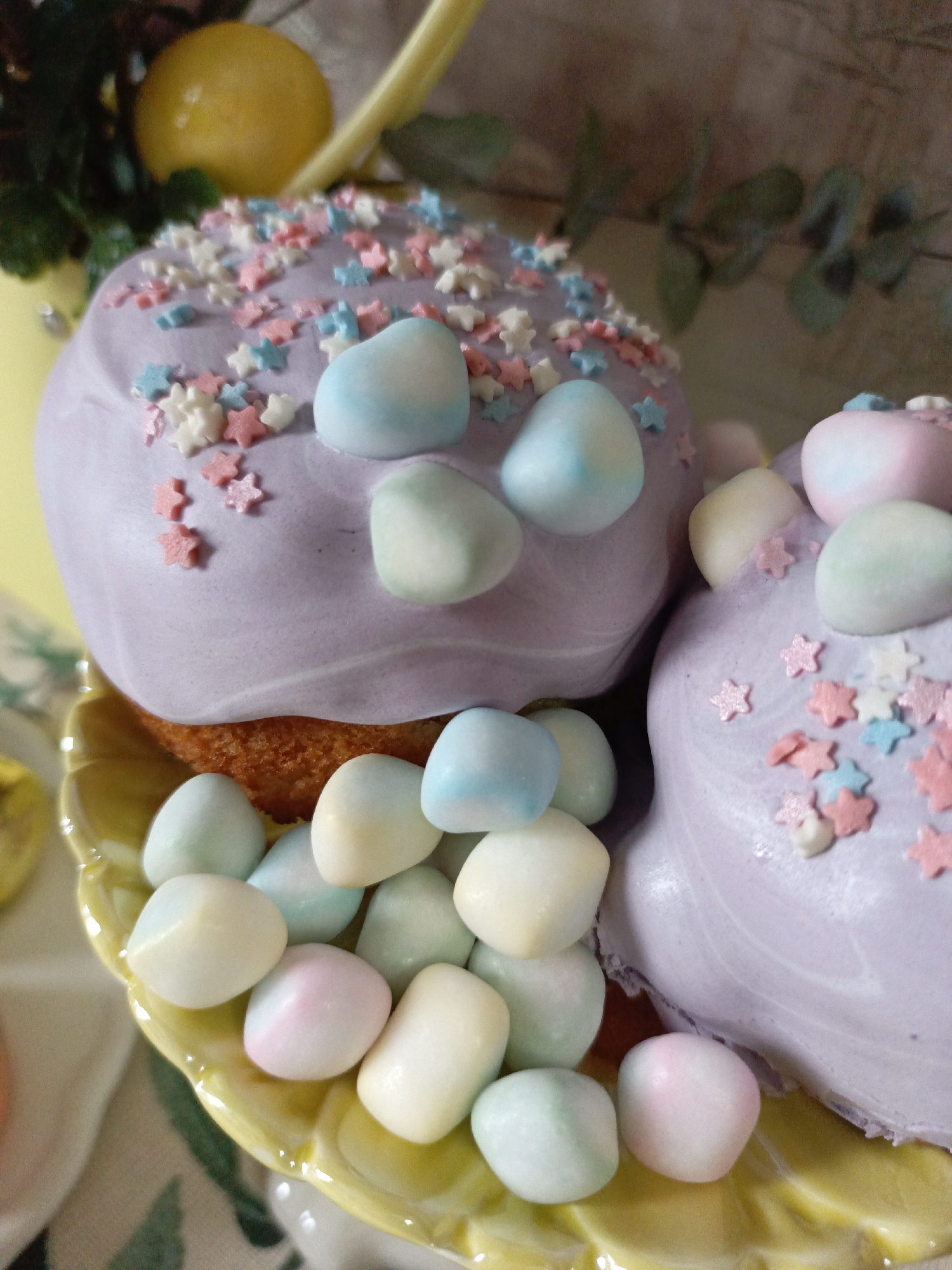 The image shows two cupcakes on a yellow plate. The cupcakes are topped with light purple frosting and decorated with pastel-colored star sprinkles and teardrop-shaped candies. The plate also contains a number of the teardrop candies. The background includes a lemon and some greenery.