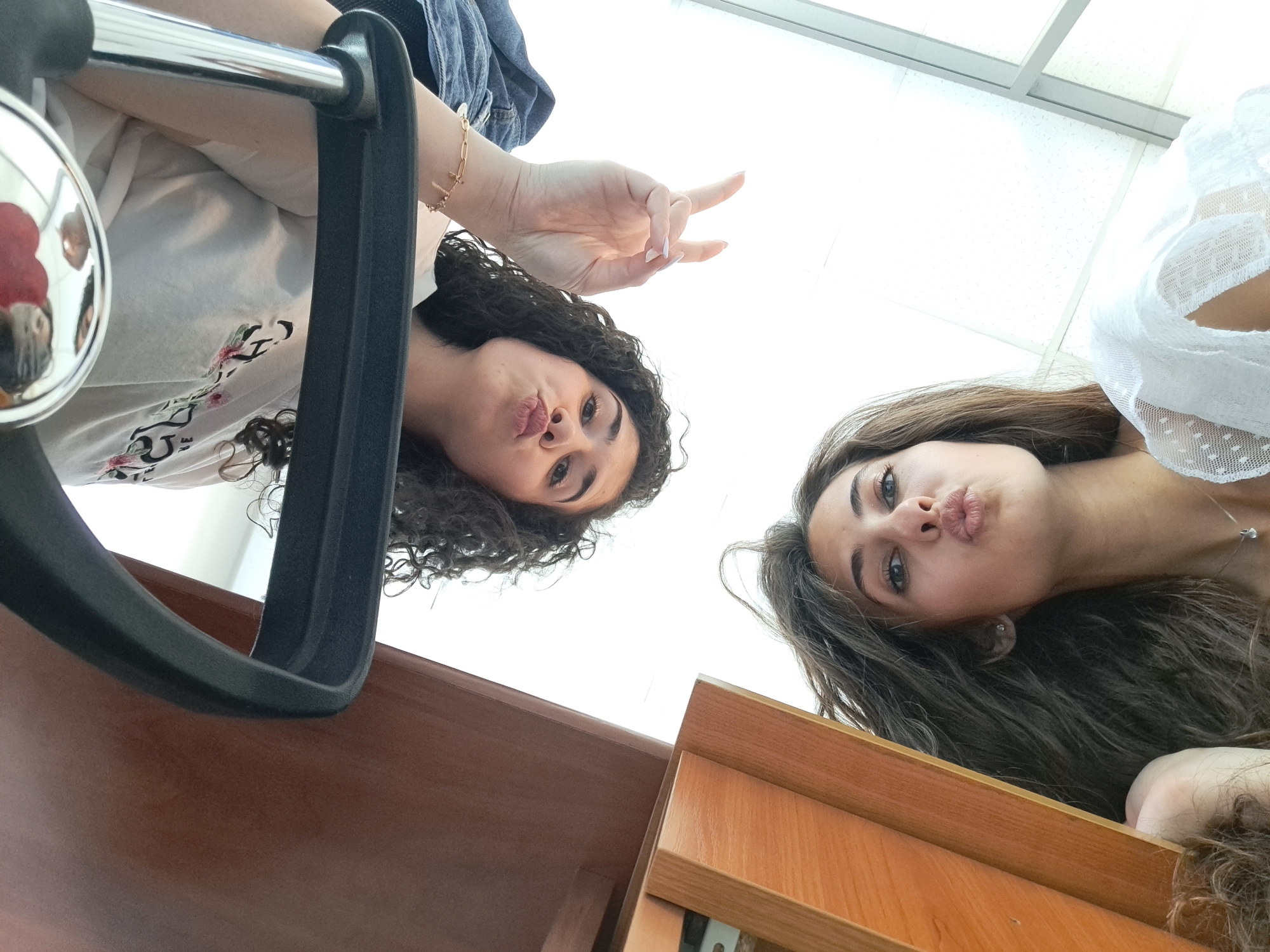 The image is a low-angle selfie featuring two young women. The woman on the left has dark, curly hair and is wearing a light-colored t-shirt with a floral design. Her hand is raised, displaying a "peace" sign. She is sitting in a black office chair. The woman on the right has long, brown hair and is wearing a white top with a delicate pattern. Both women are pursing their lips as if posing for a kiss. The background is a bright, white surface, likely a ceiling. A wooden desk or cabinet is visible in the lower portion of the frame. The location in the city of Sofia, Bulgaria, cannot be determined from this image.